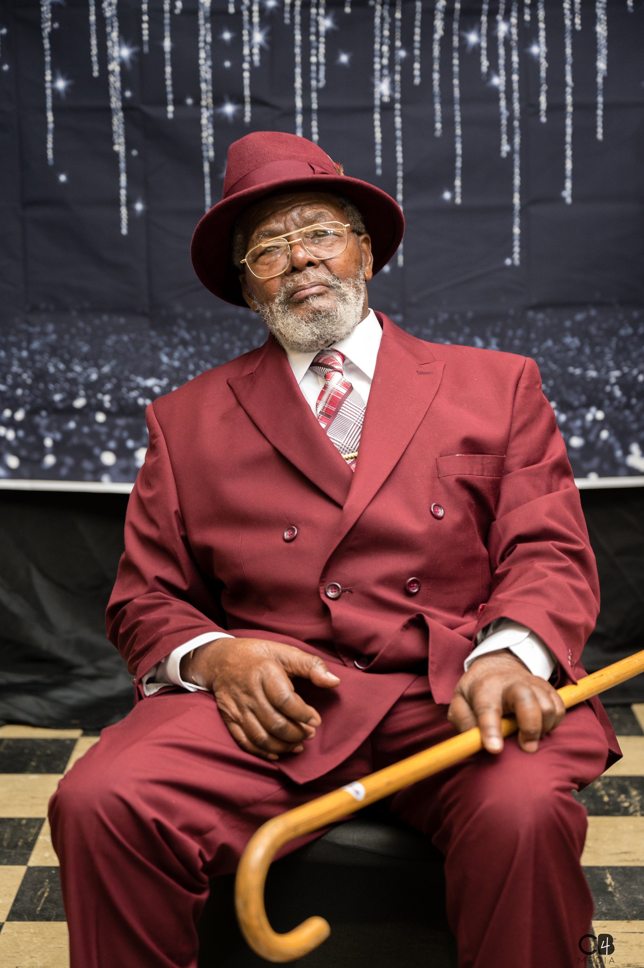 An elderly man dressed in a burgundy suit with a matching hat and tie, sitting on a chair holding a cane, with a black backdrop decorated with silver hanging beads.