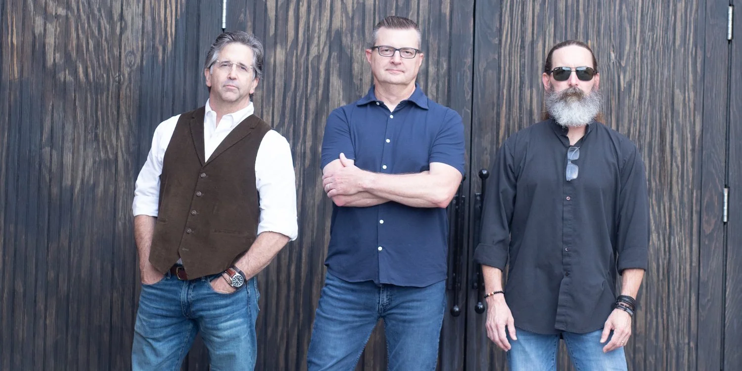 Three men standing side by side against a wooden wall, with the middle man crossing his arms and the other two hands in pockets, wearing casual shirts and jeans, one with sunglasses and a beard, one with glasses, and one with a watch.