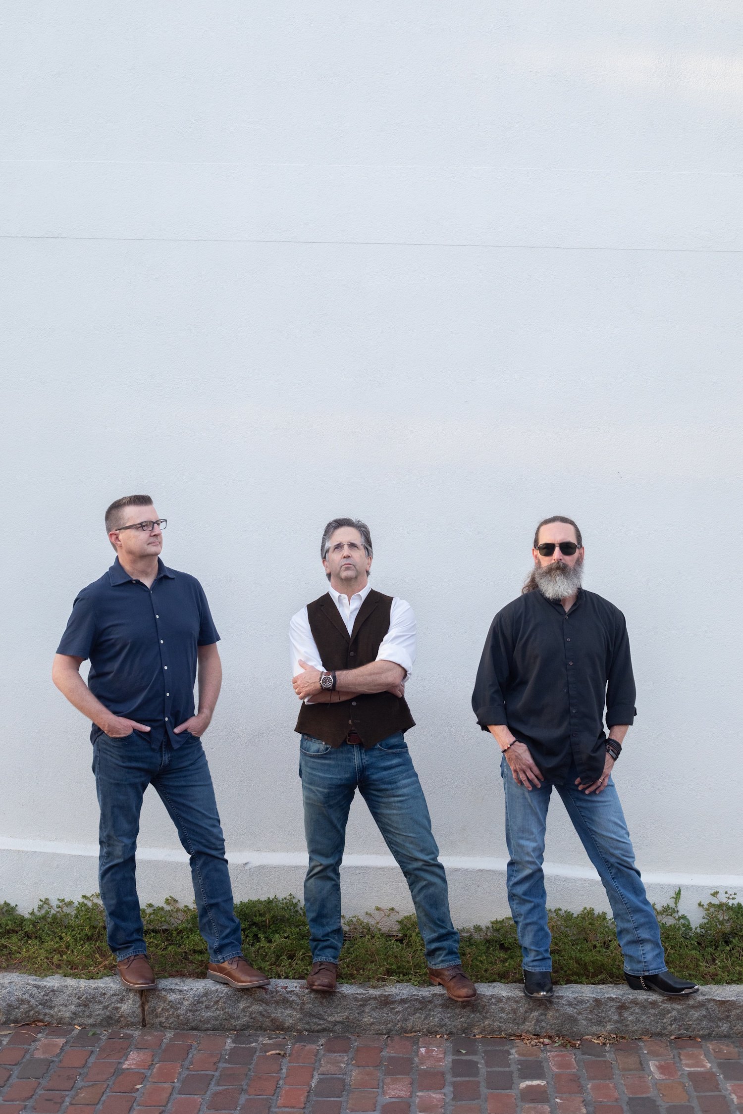 Three men standing outside against a plain white wall with brick pavement and some grass at the base, posing for a photo.