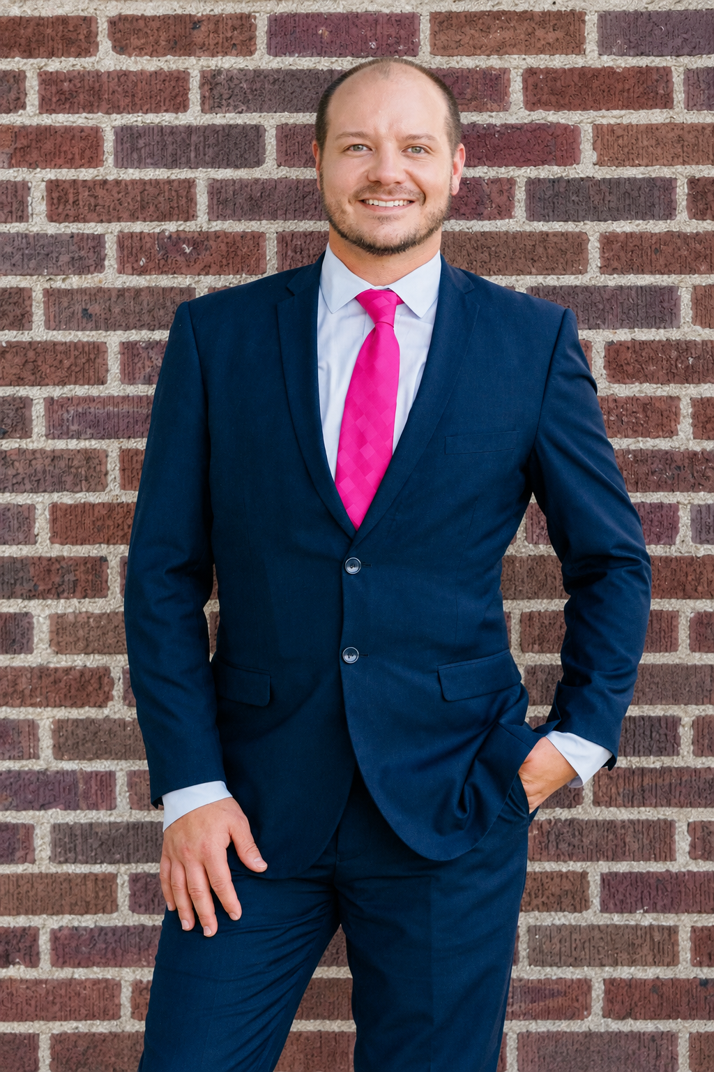Man in a navy suit with a pink tie, standing outdoors near a building, smiling.