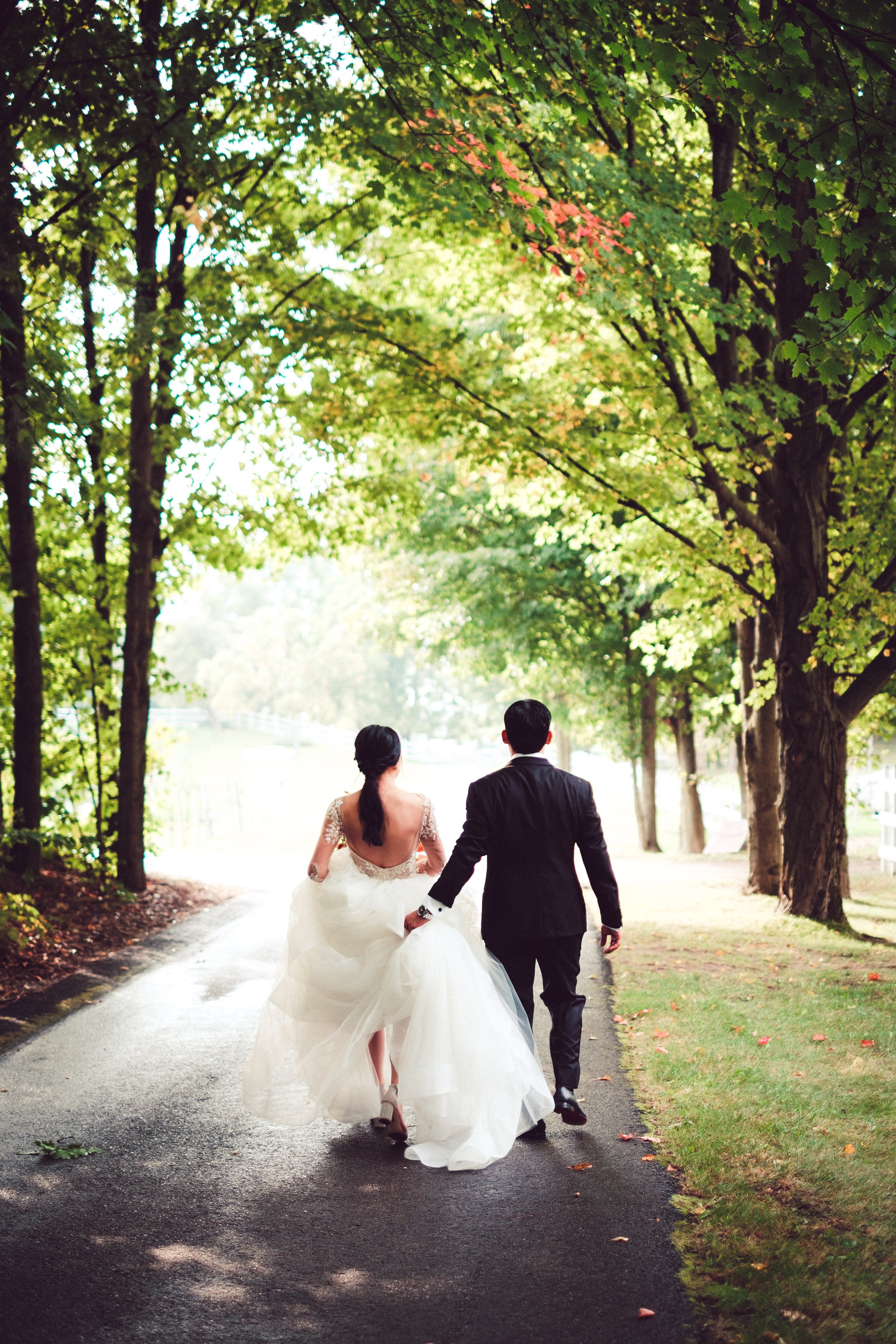 Ponytail bride walking on tree lined path groom holding dress