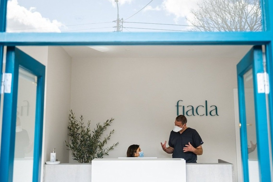 A nurse or medical professional talking to a patient at a reception desk in a modern medical office called 'fiadle'. Both are wearing masks.