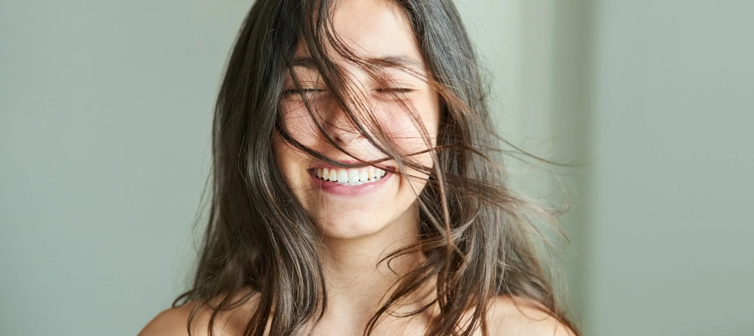A young woman with long, wavy brown hair laughing with her eyes closed, against a light, neutral background.