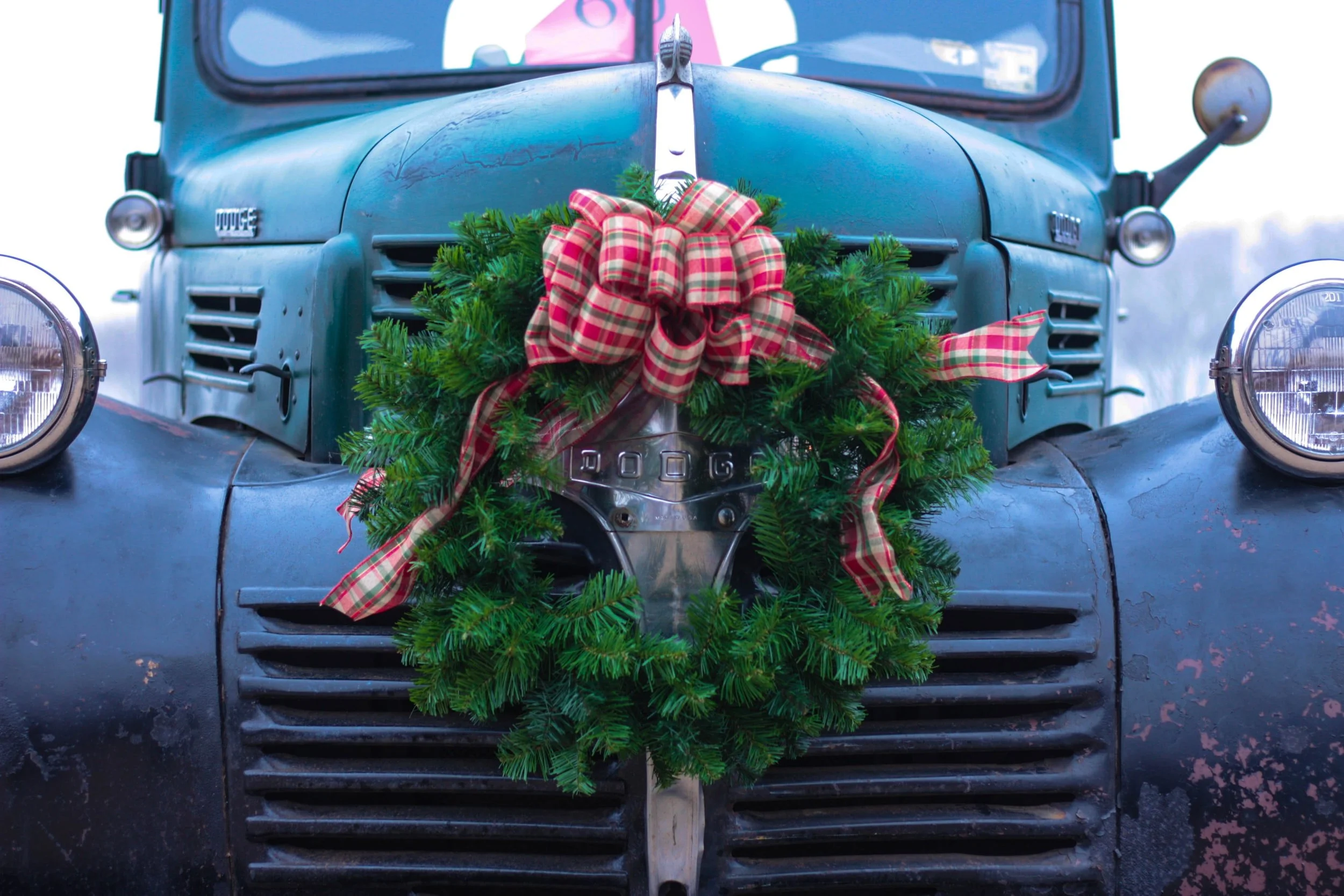 A vintage green Dodge truck decorated with a Christmas wreath featuring a red plaid ribbon on the front grille.