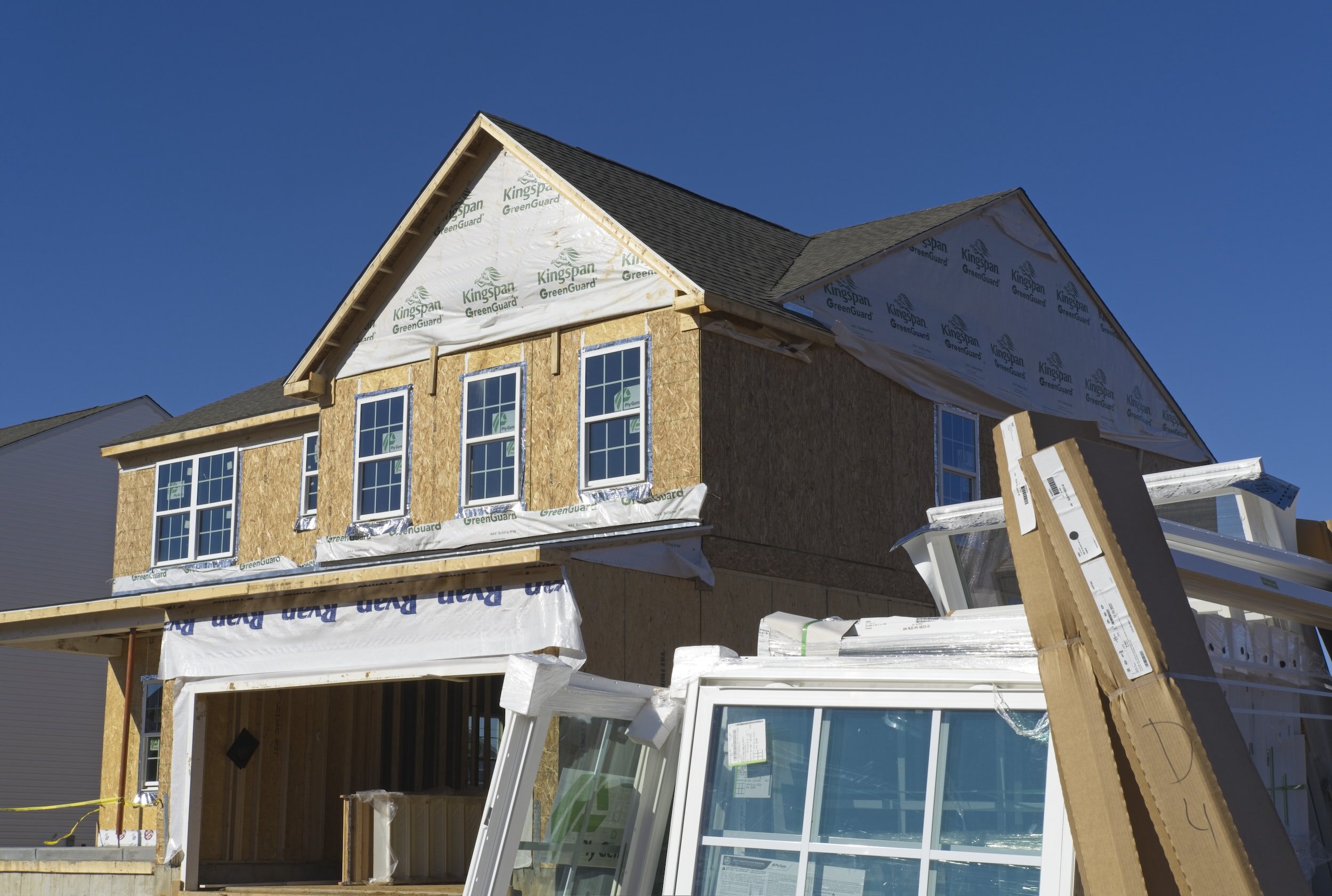 House under construction with exposed wooden frames and wrapped in building materials, windows stacked in foreground.