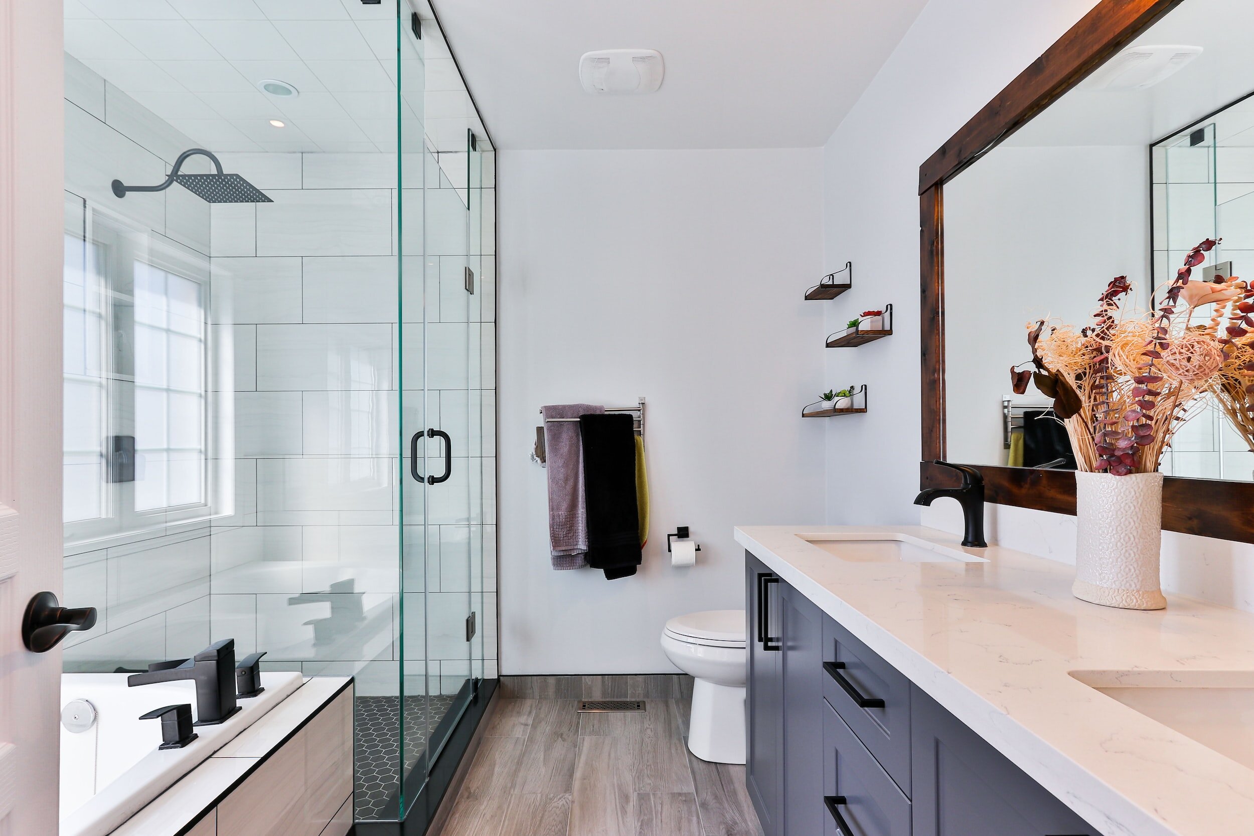 Modern bathroom with glass shower, bathtub, wooden floor, double vanity with white countertop, large mirror, and decorative vase.