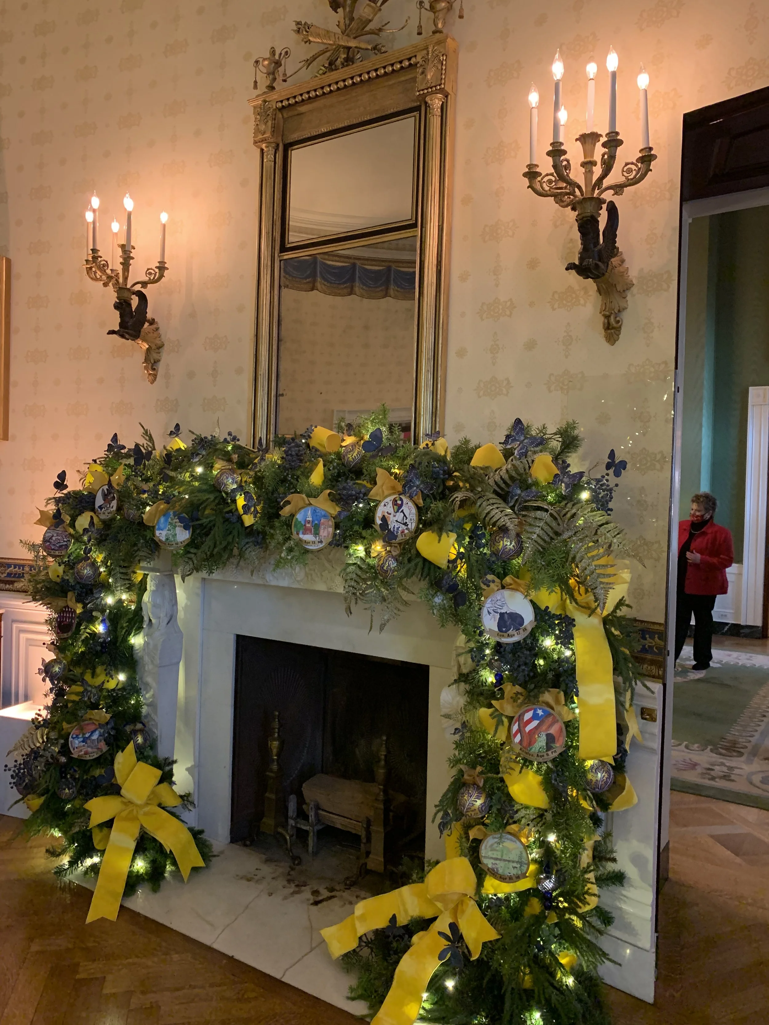 Elegant living room fireplace decorated with lush evergreen garland, yellow bows, and ornaments featuring various images. The room includes a large mirror above the mantel, flanked by decorative wall sconces.