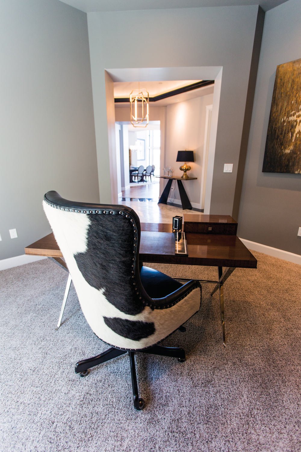 Modern office space with a cowhide chair, dark wooden desk, and beige carpet. The room opens into a hallway with a dining area in the background, featuring a chandelier and a lit lamp on a table.