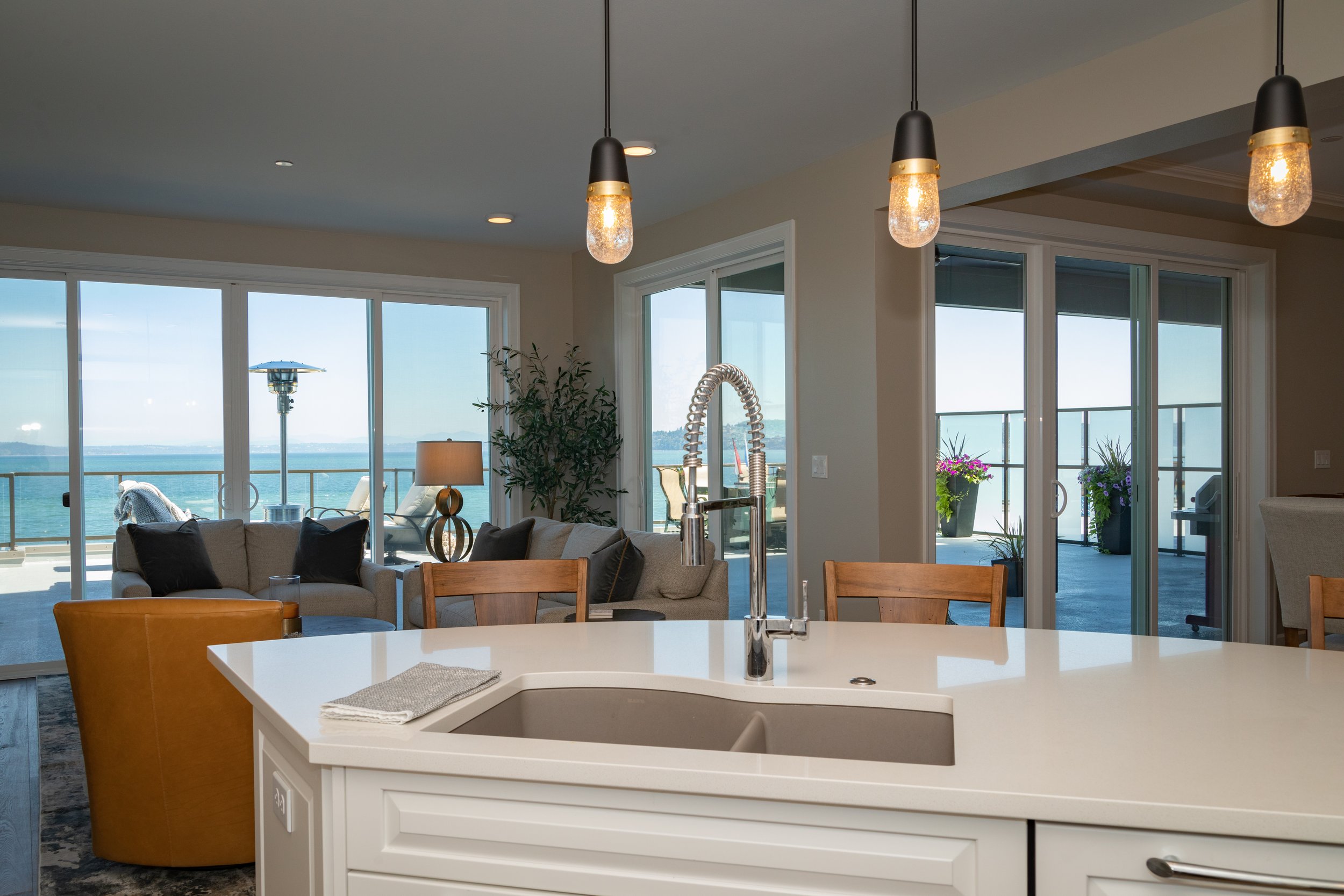 Modern kitchen and living room with waterfront view, featuring a white island countertop, stainless steel faucet, pendant lights, and sliding glass doors leading to a patio.
