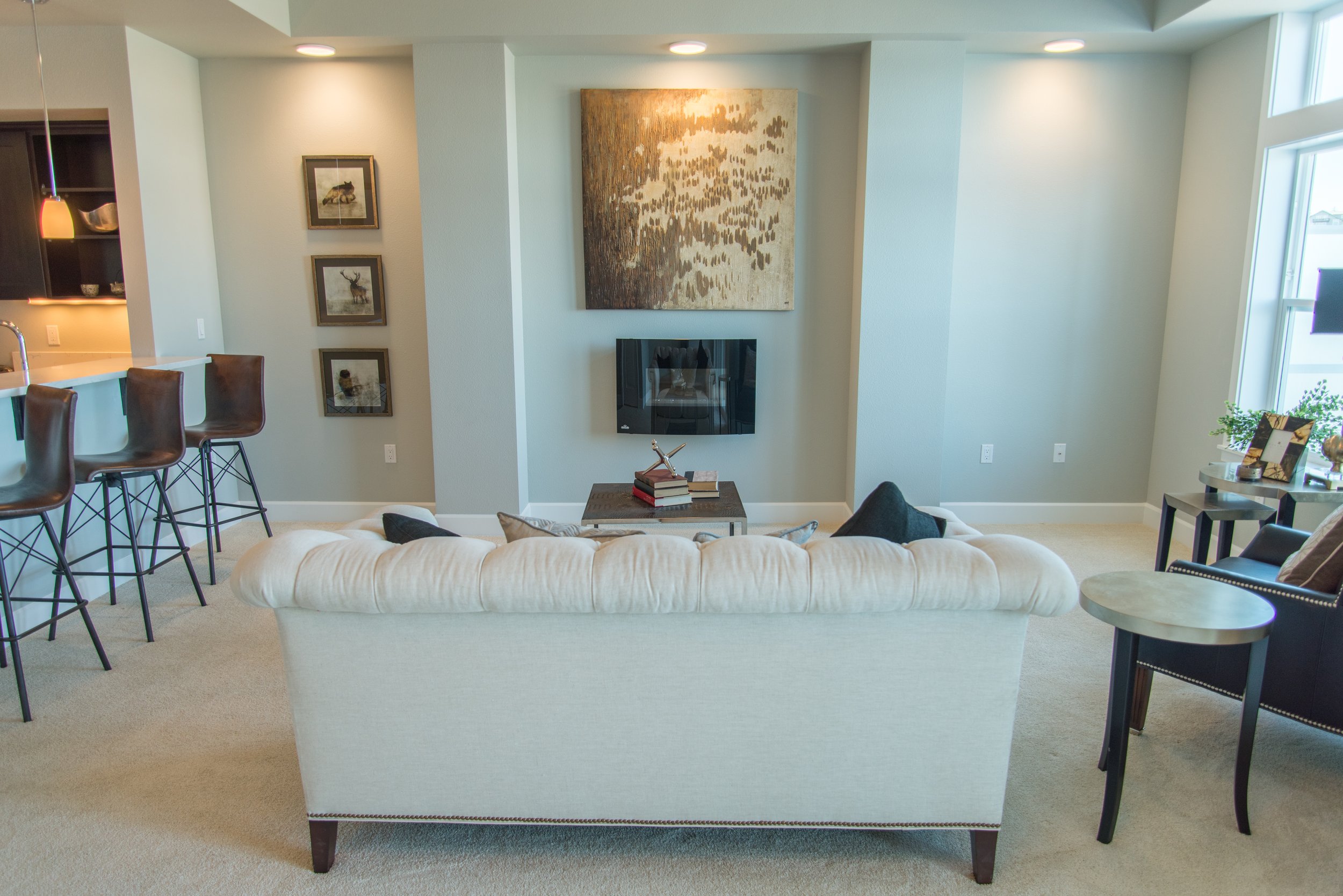 Modern living room with a beige sofa, abstract wall art, bar stools, and a small side table with books.
