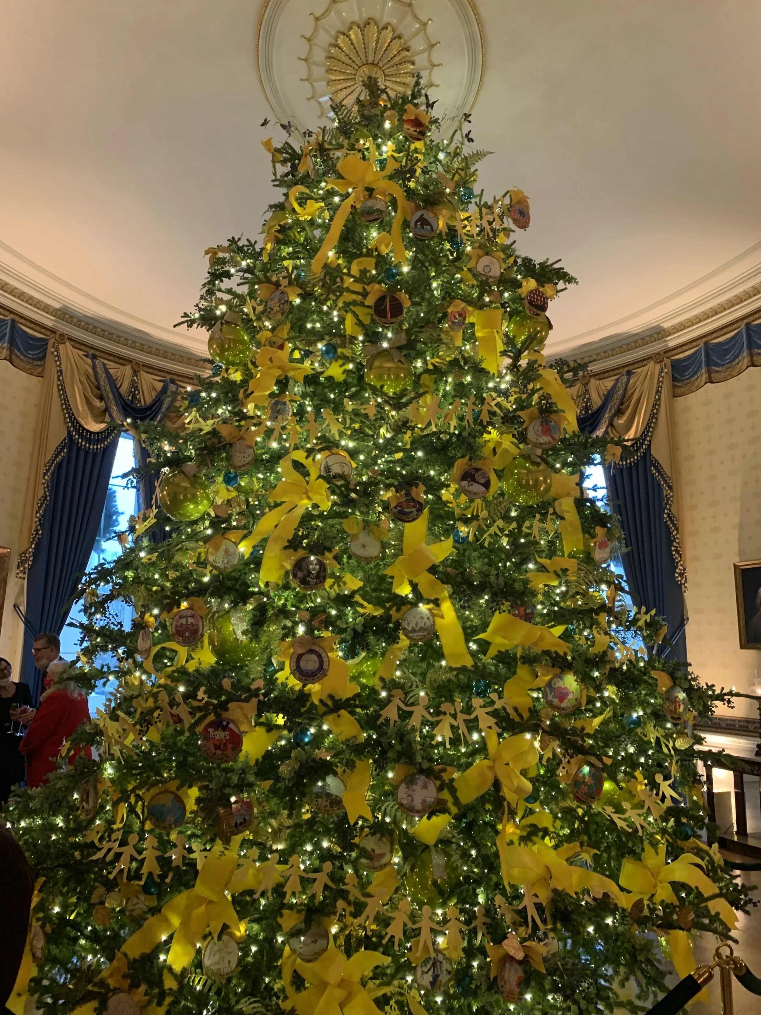 Christmas tree decorated with yellow ribbons, ornaments, and lights in a luxurious room with blue curtains.