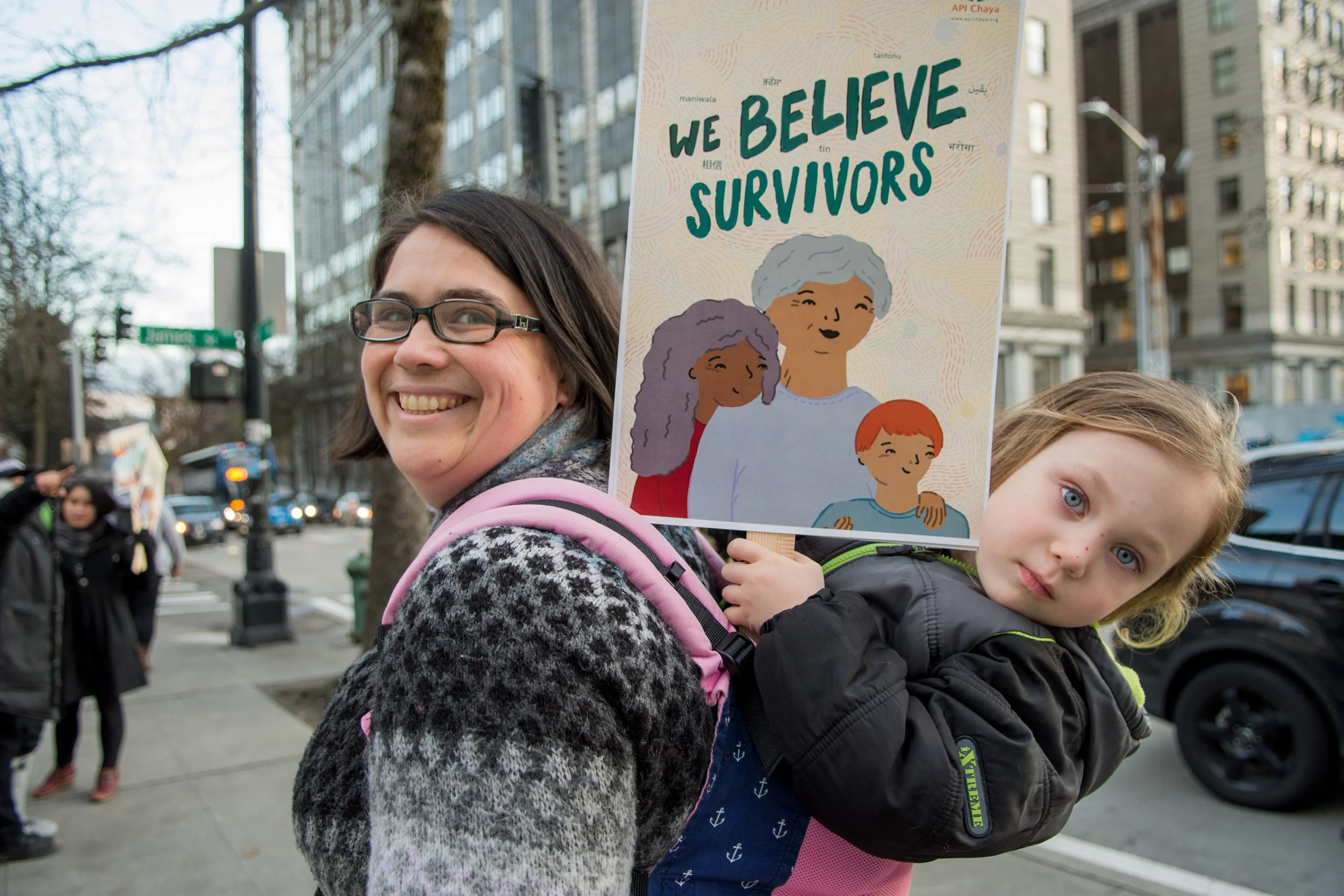 A parent and child on the sidewalk in the city. The child is holding an API Chaya sign that reads, “We Believe Survivors”