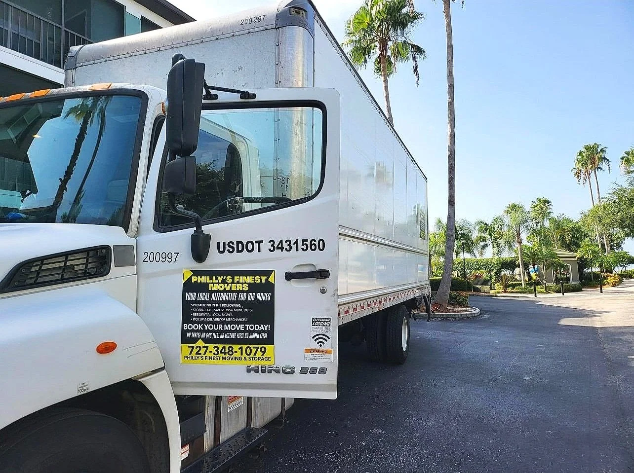 A white moving truck with black and yellow signage parked outside on a sunny day, surrounded by palm trees and greenery.