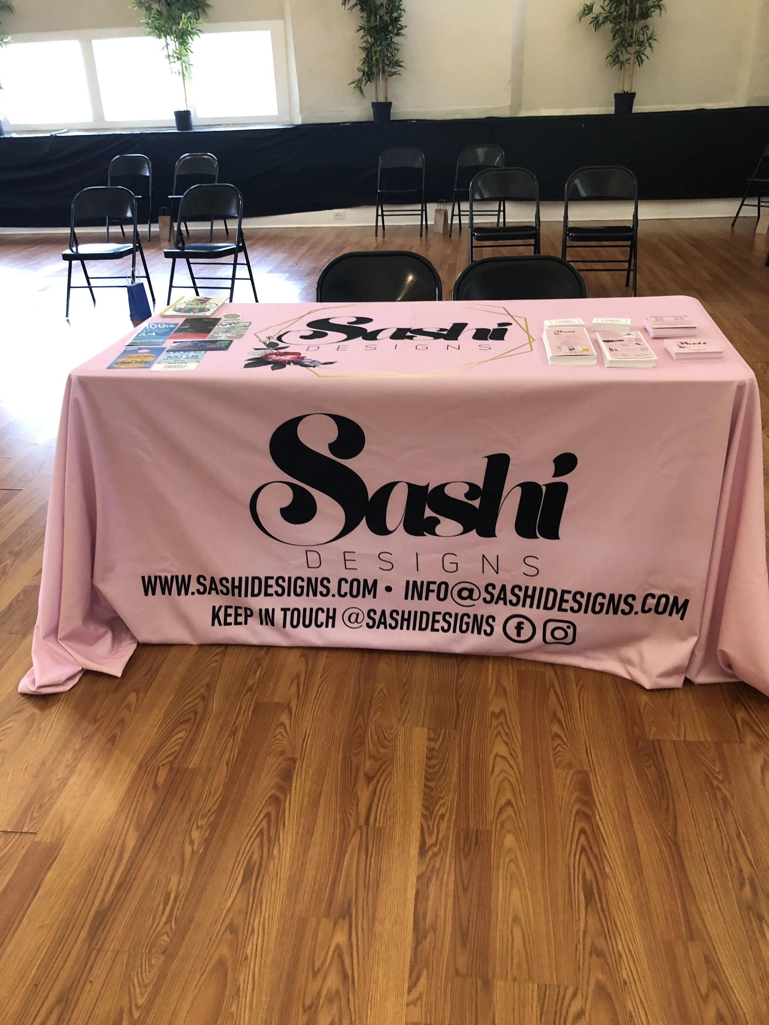 A table with a pink tablecloth featuring the logo and contact information for Sashi Designs. The table has pamphlets and flyers on it, with chairs arranged behind and beside it in a room with wooden flooring and large windows in the background.