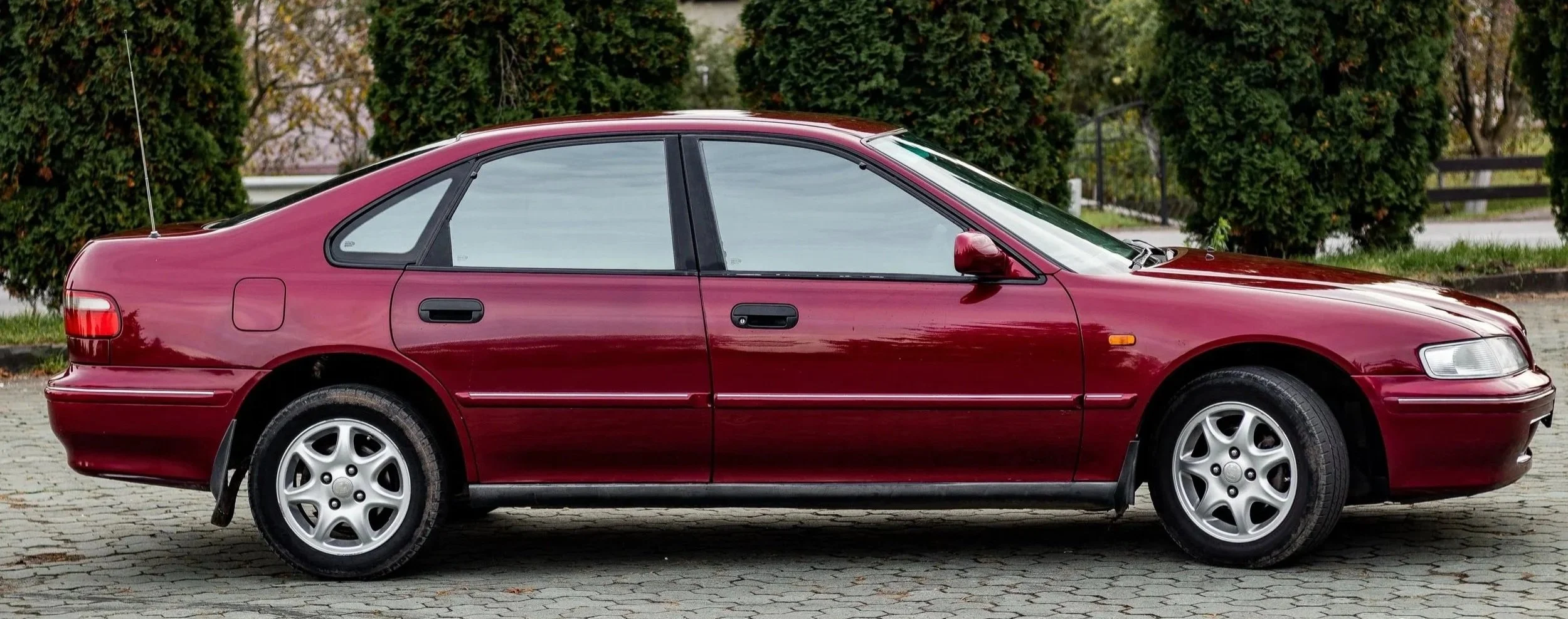Side view of a maroon sedan parked on a cobblestone driveway with trees and greenery in the background.