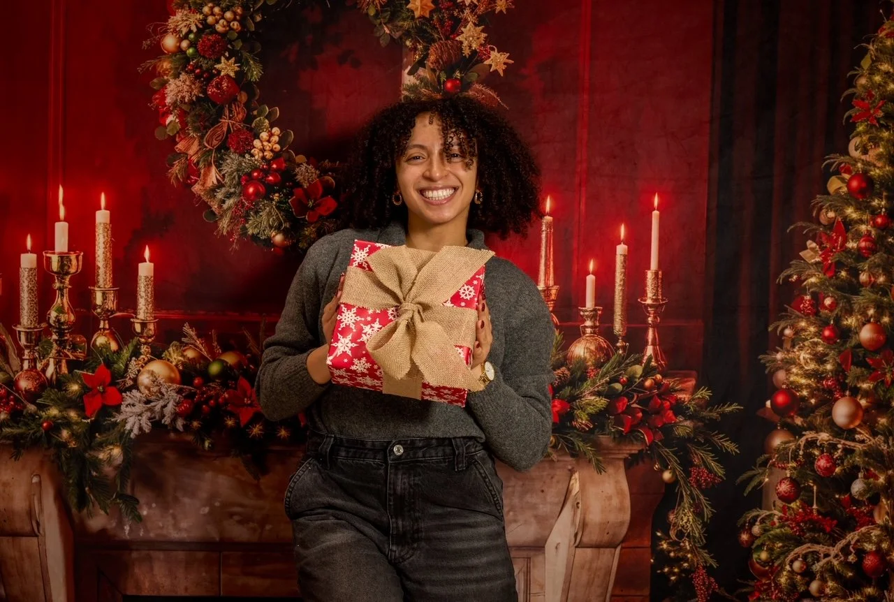Person holding a wrapped gift in front of a festive Christmas background with candles and a wreath.
