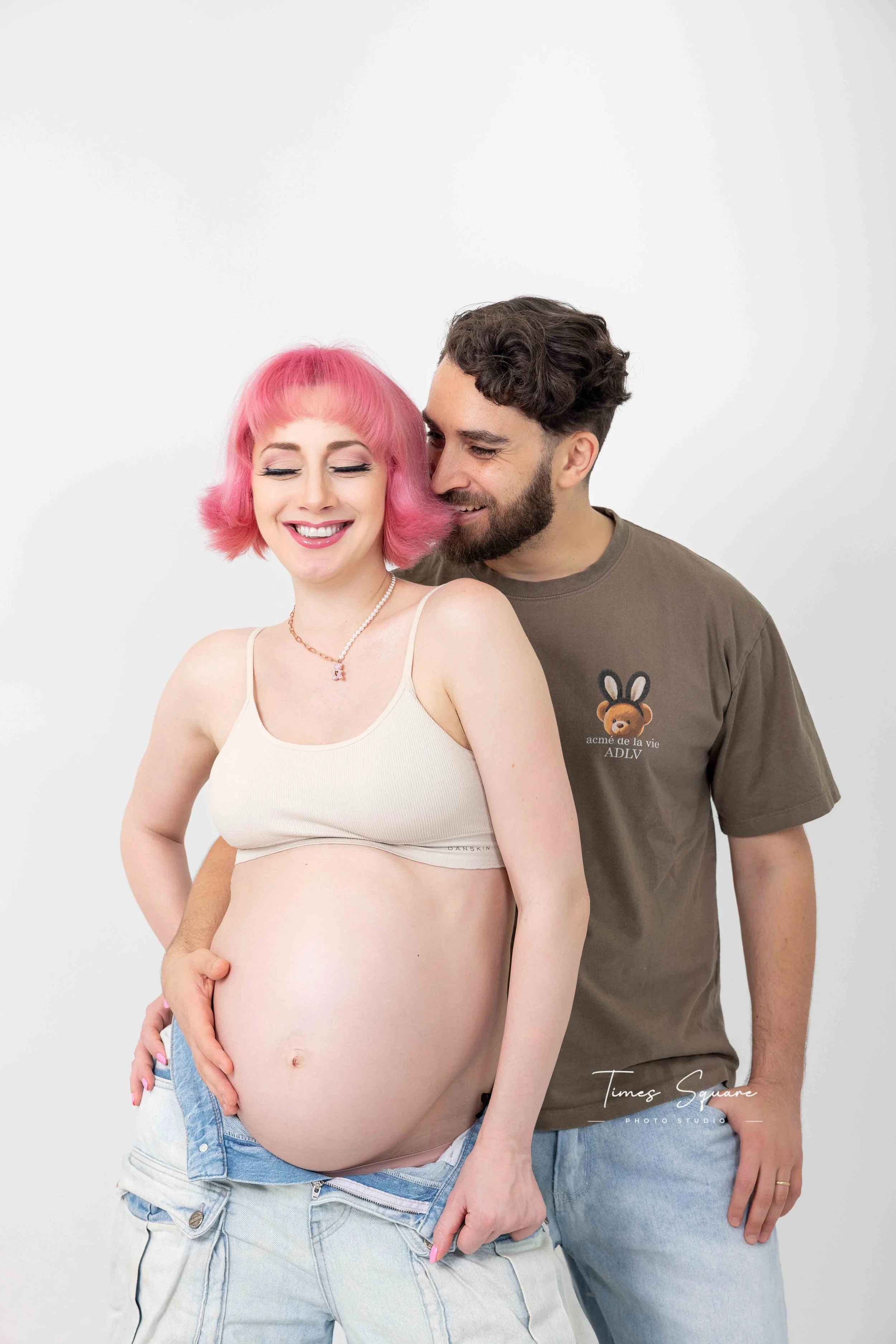 A pregnant woman with pink hair smiling with eyes closed, wearing a cream tank top, light denim shorts, and a pearl necklace, standing next to a man with dark curly hair and beard, who is wearing a gray T-shirt with a bear graphic and text, and smiling at her against a plain white background.