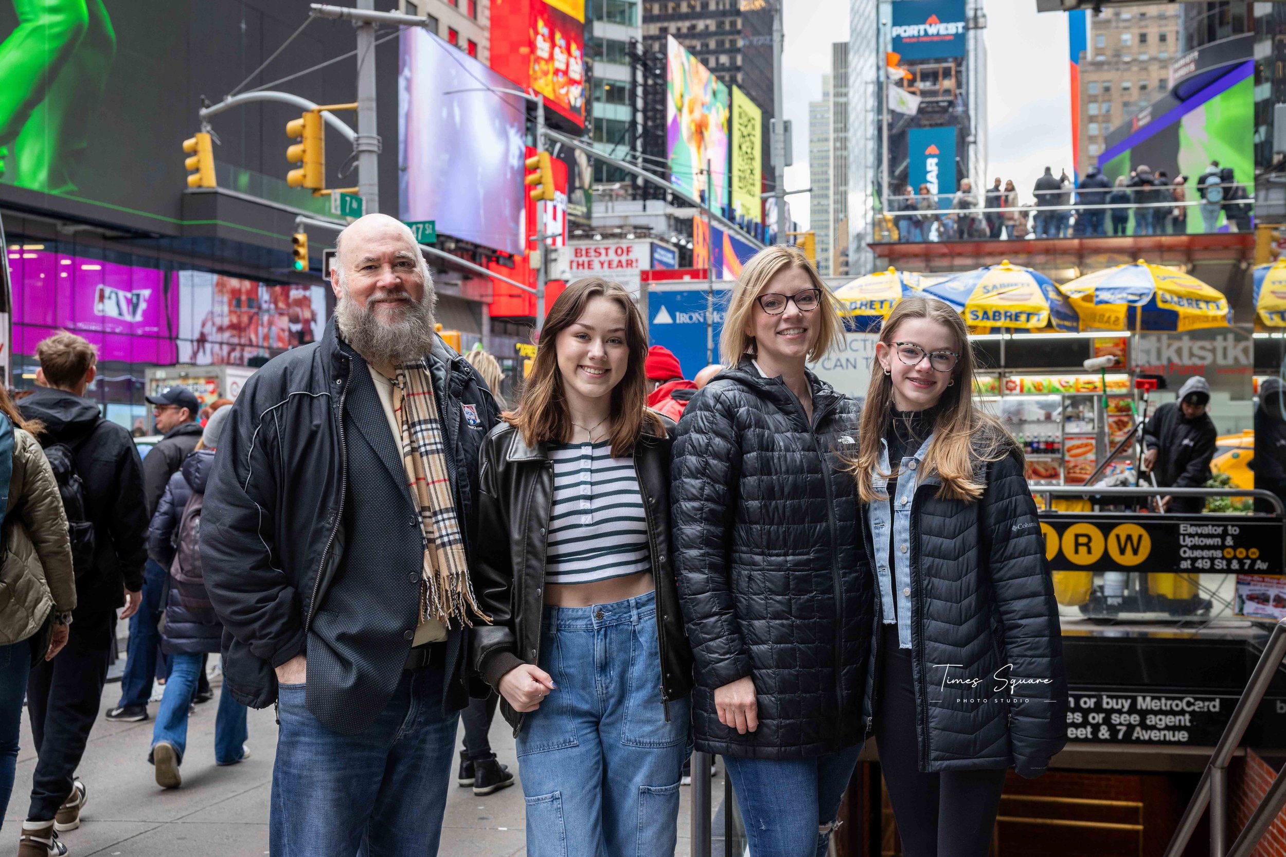A family of four posing in Times Square, New York City, with colorful billboards and bright lights in the background. They are dressed in winter clothing with three women and one man, standing next to a subway entrance and a Sabrett hot dog stand.