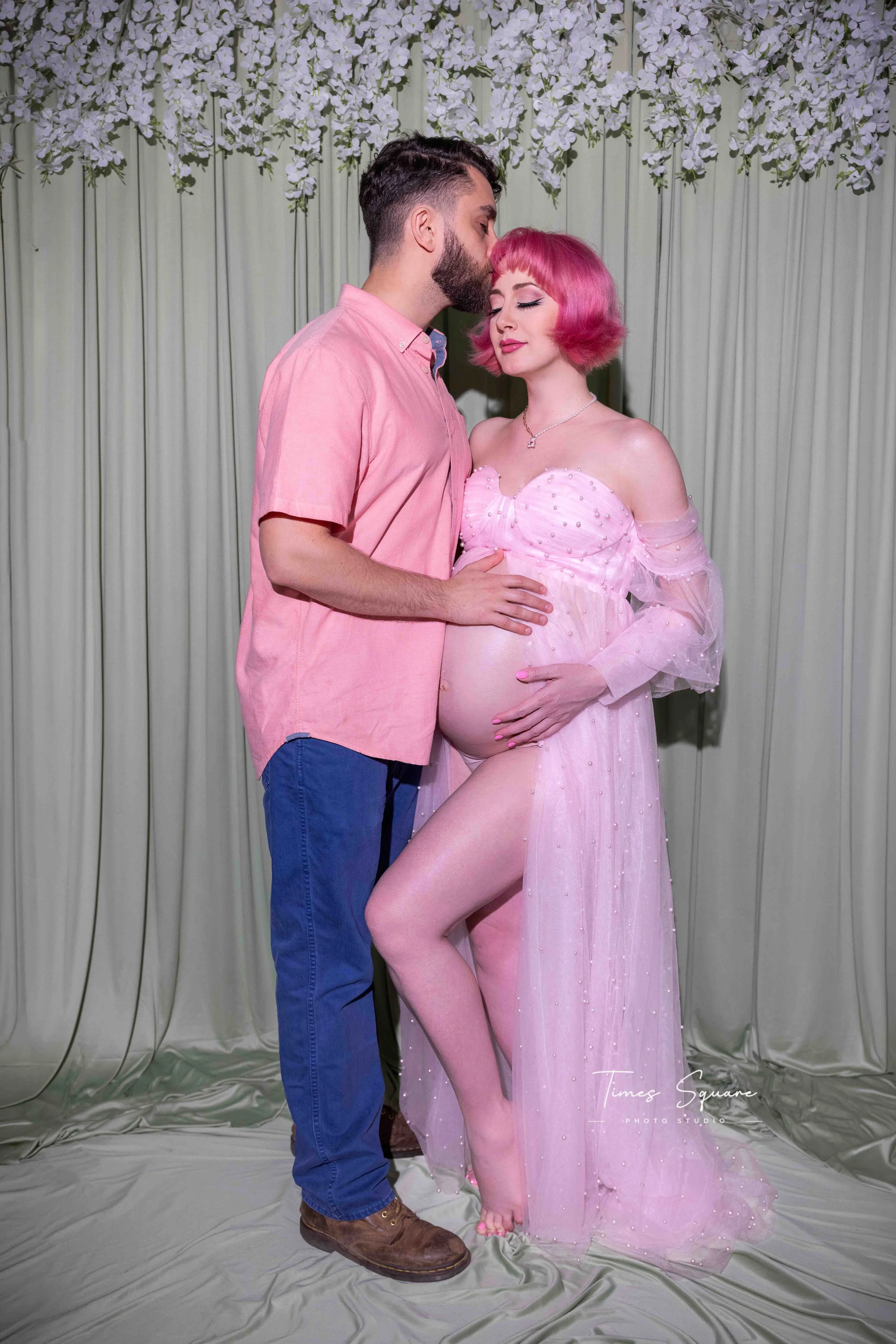 A man with dark hair, beard, wearing a pink shirt and blue jeans, is kissing a woman with vibrant pink hair, wearing a sheer pink dress, in front of a floral backdrop.