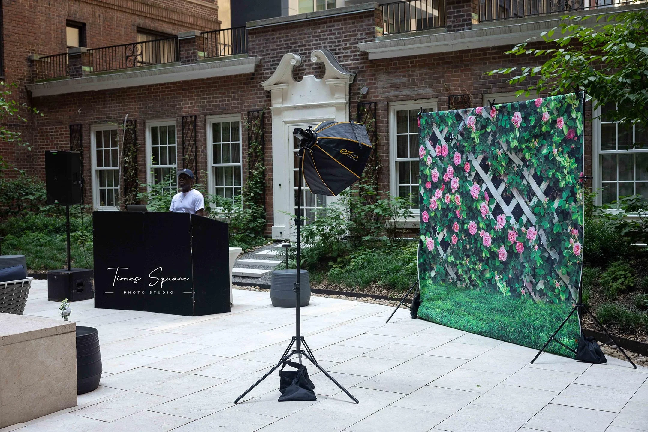 Outdoor photography setup with a floral backdrop, lighting equipment, and a person behind a podium labeled "Times Square Photo Studio" in a courtyard.