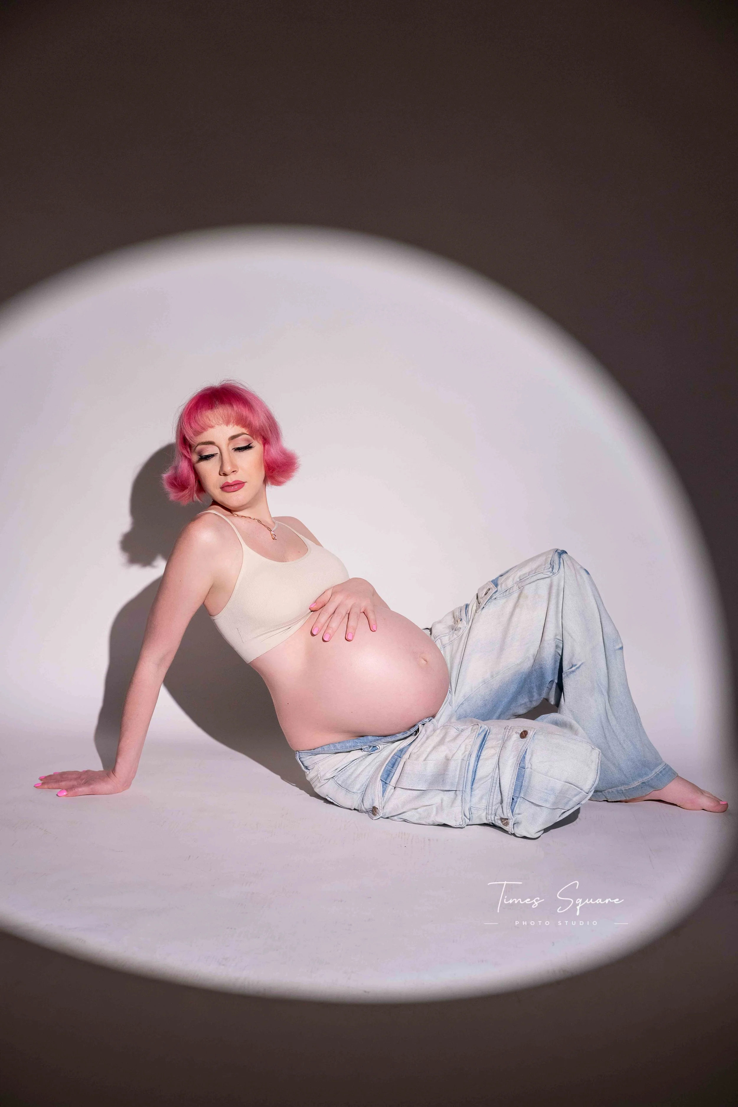 A woman with pink hair and makeup sitting on the floor with one arm supporting her. She is pregnant, wearing a cream tank top and light blue jeans, with a white background and a circular spotlight effect.