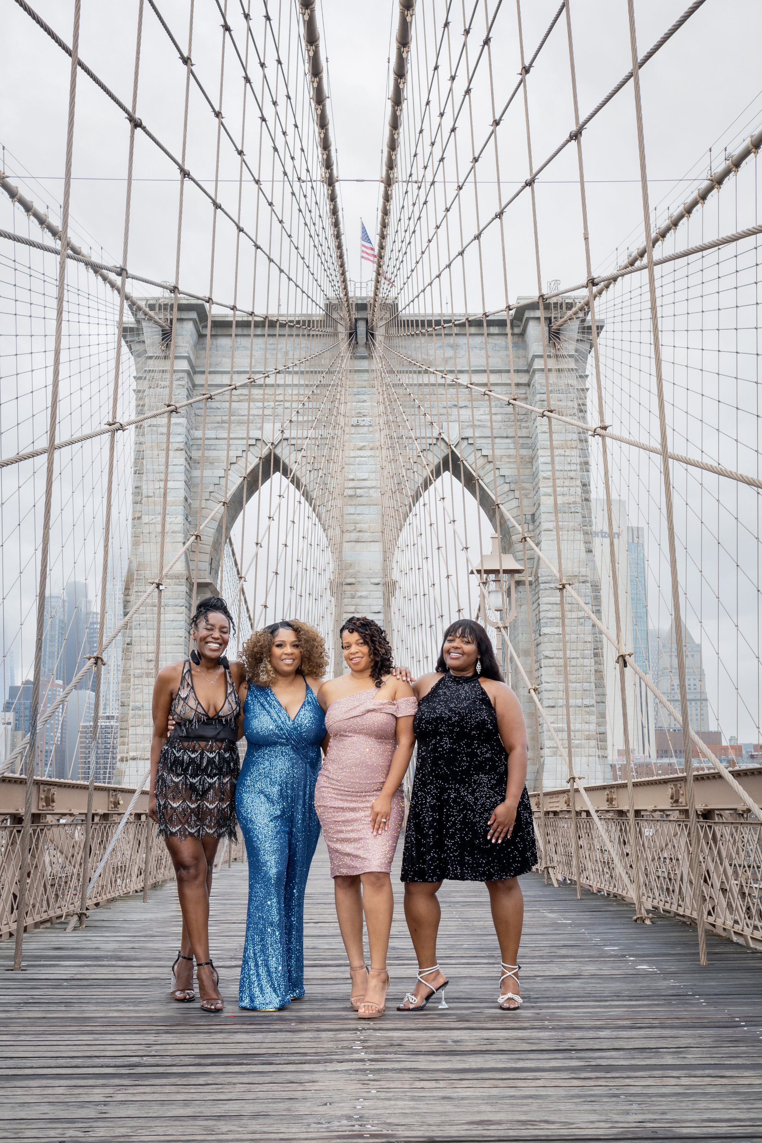 Four women wearing formal dresses posing on the Brooklyn Bridge.