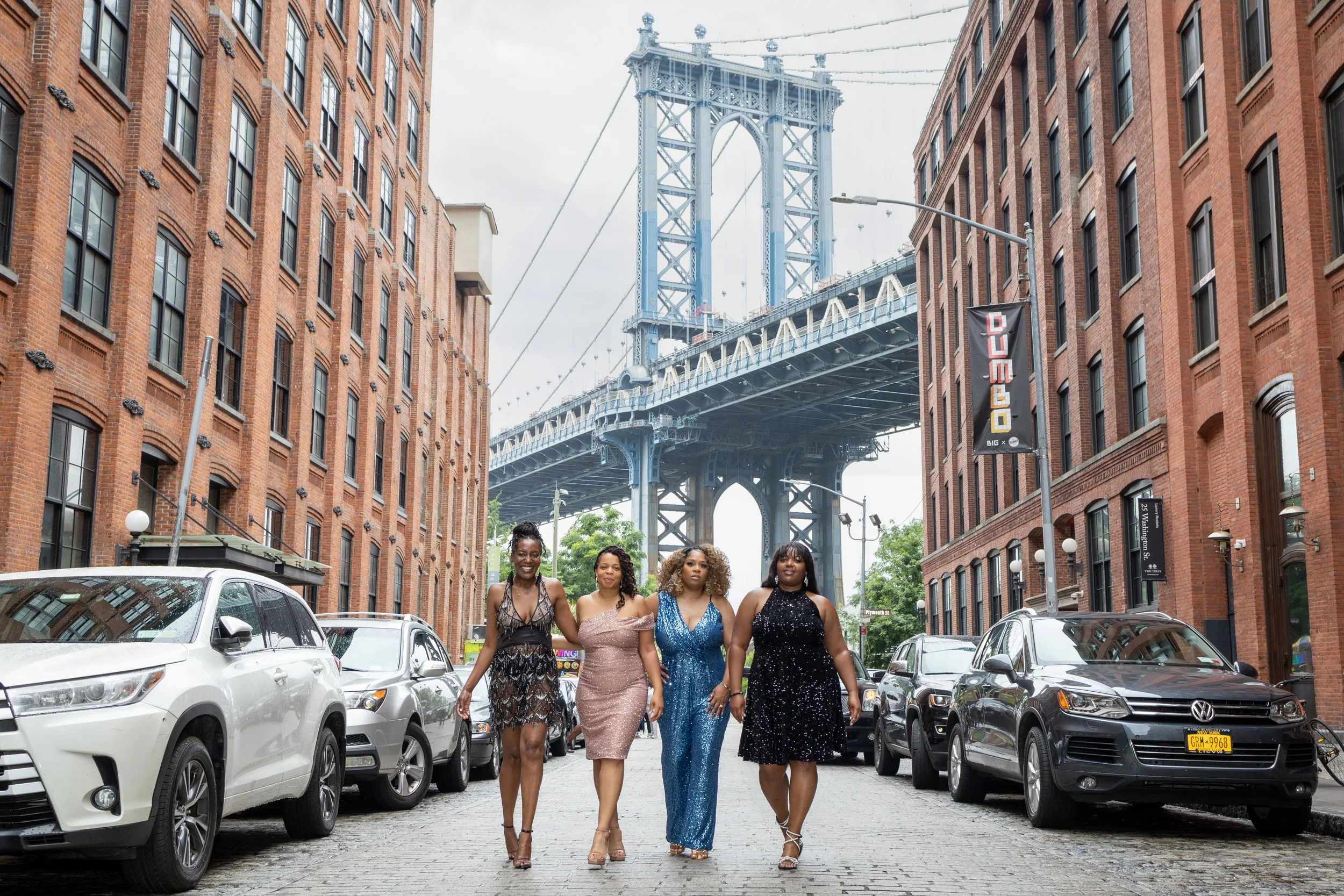 Four women in formal dresses walking on a cobblestone street in Dumbo, Brooklyn, with the Manhattan Bridge in the background.
