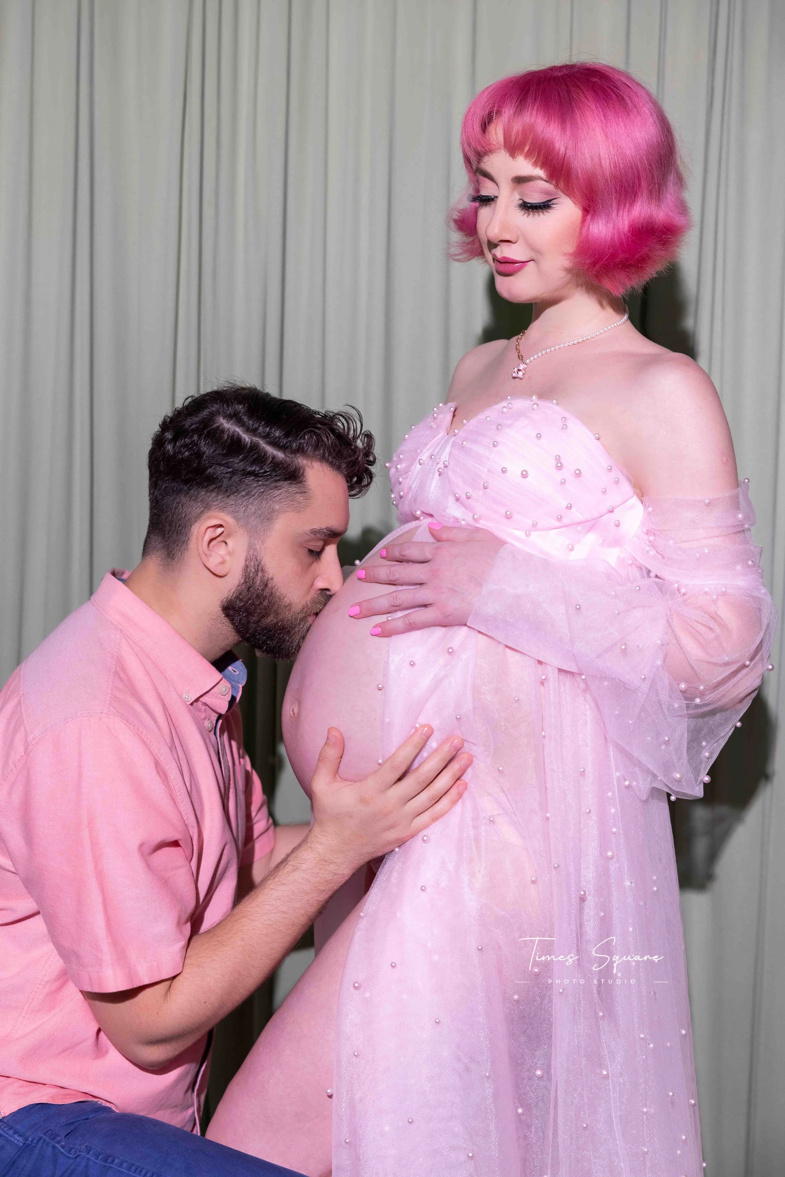 A man kneeling and kissing a pregnant woman's belly while she stands, wearing a pink dress with pearl embellishments, in front of a cream-colored curtain.