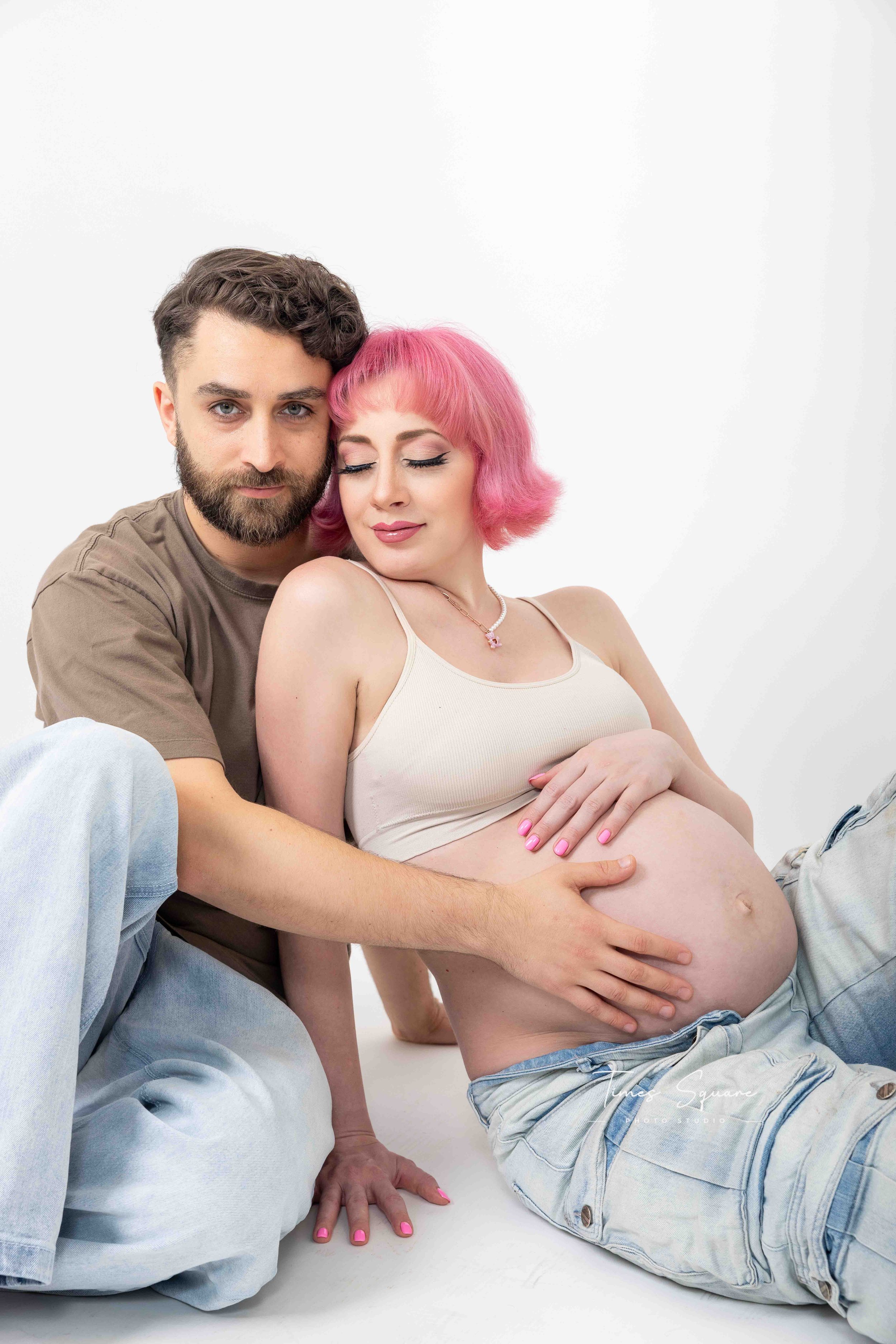 A pregnant woman with pink hair and a beige tank top sitting against a white background, gently holding her belly, while a man with a beard and brown hair is hugging her from the side, both with peaceful expressions.