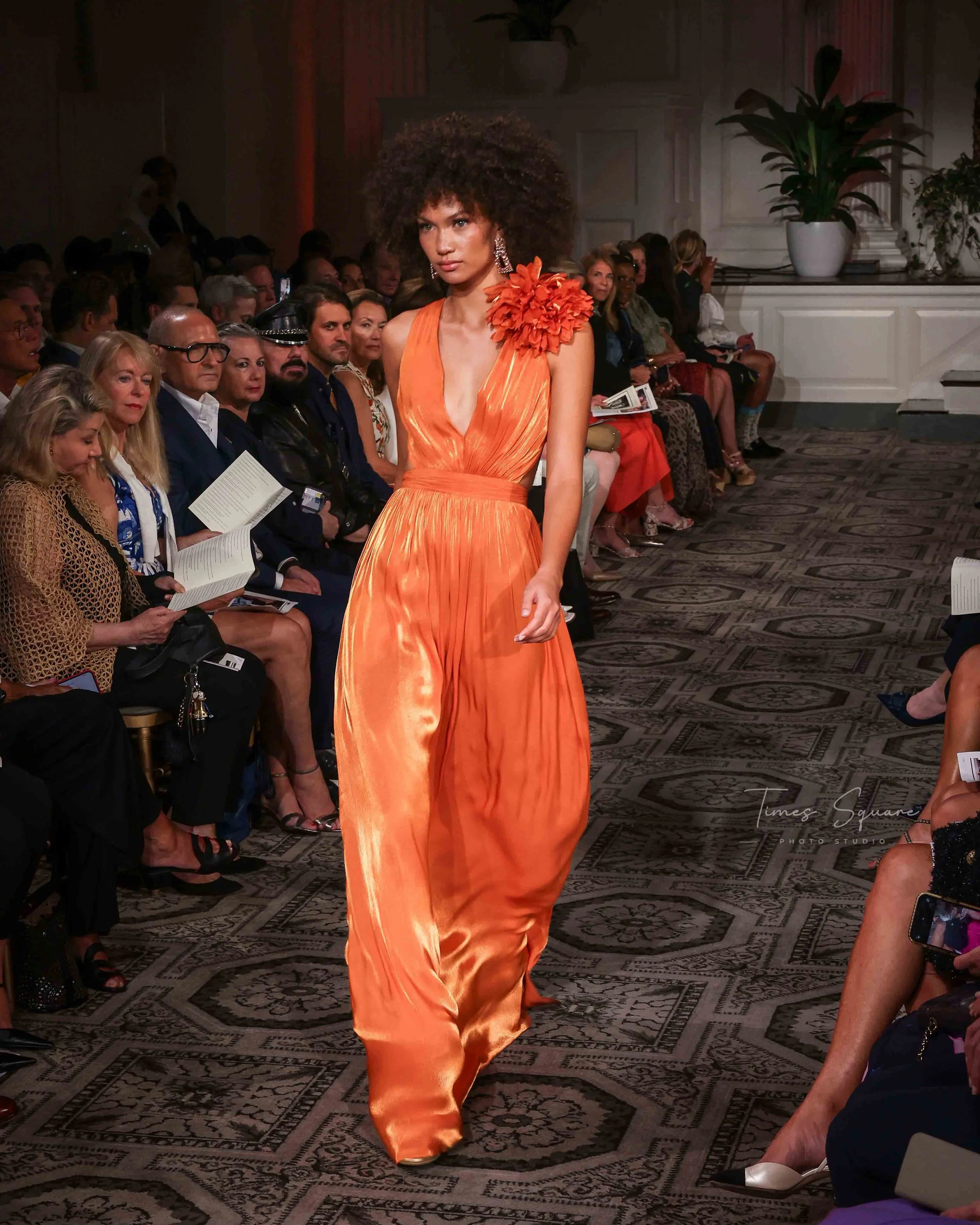 A model walks down the runway in an elegant orange gown with a flower embellishment on the shoulder, during a fashion show. The audience is seated on either side, attentively watching the event.