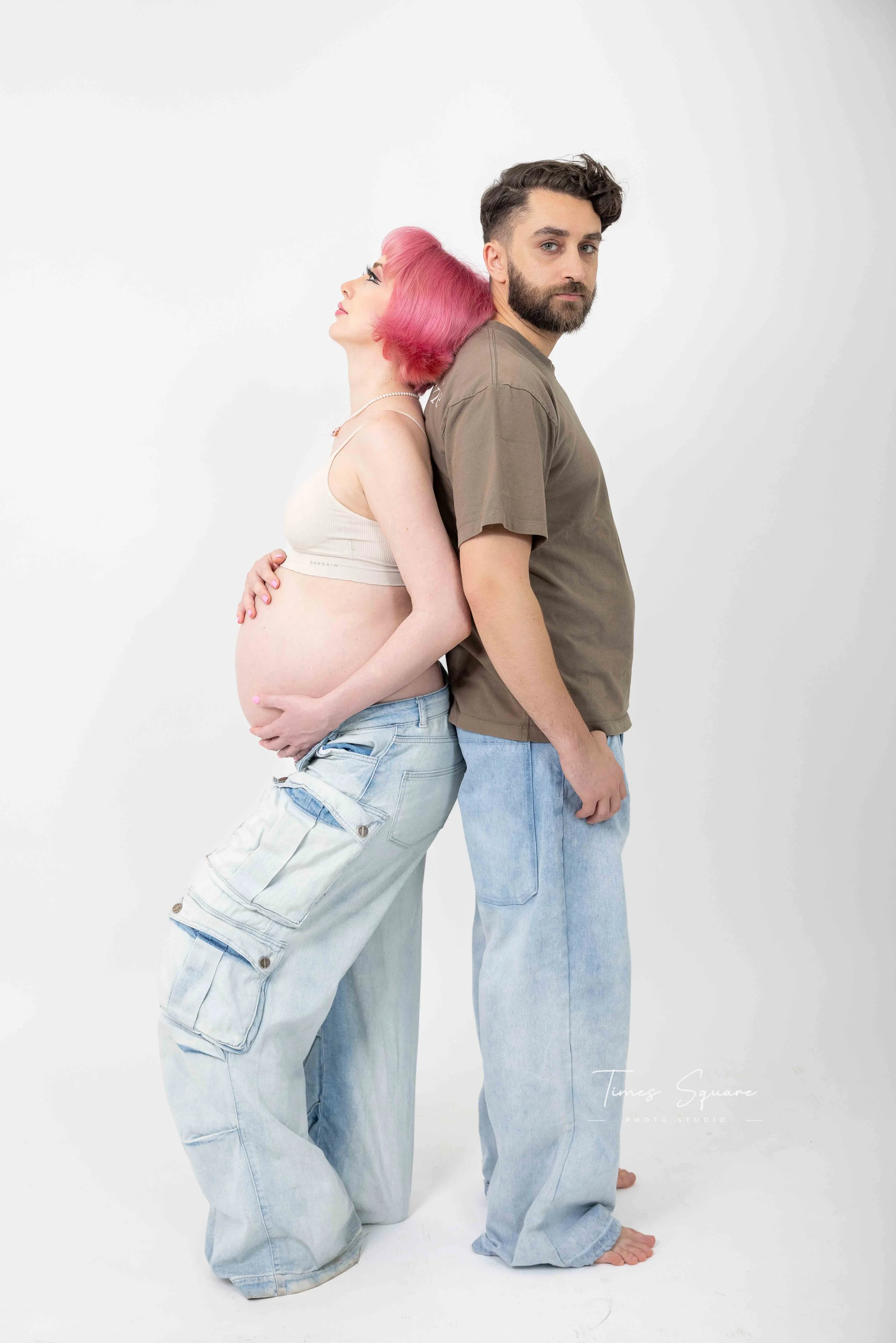 A pregnant woman with pink hair and light skin stands with her back against a man. They are both barefoot, standing on a white background, with the woman wearing a beige crop top and cargo jeans, and the man in a brown t-shirt and light blue jeans.