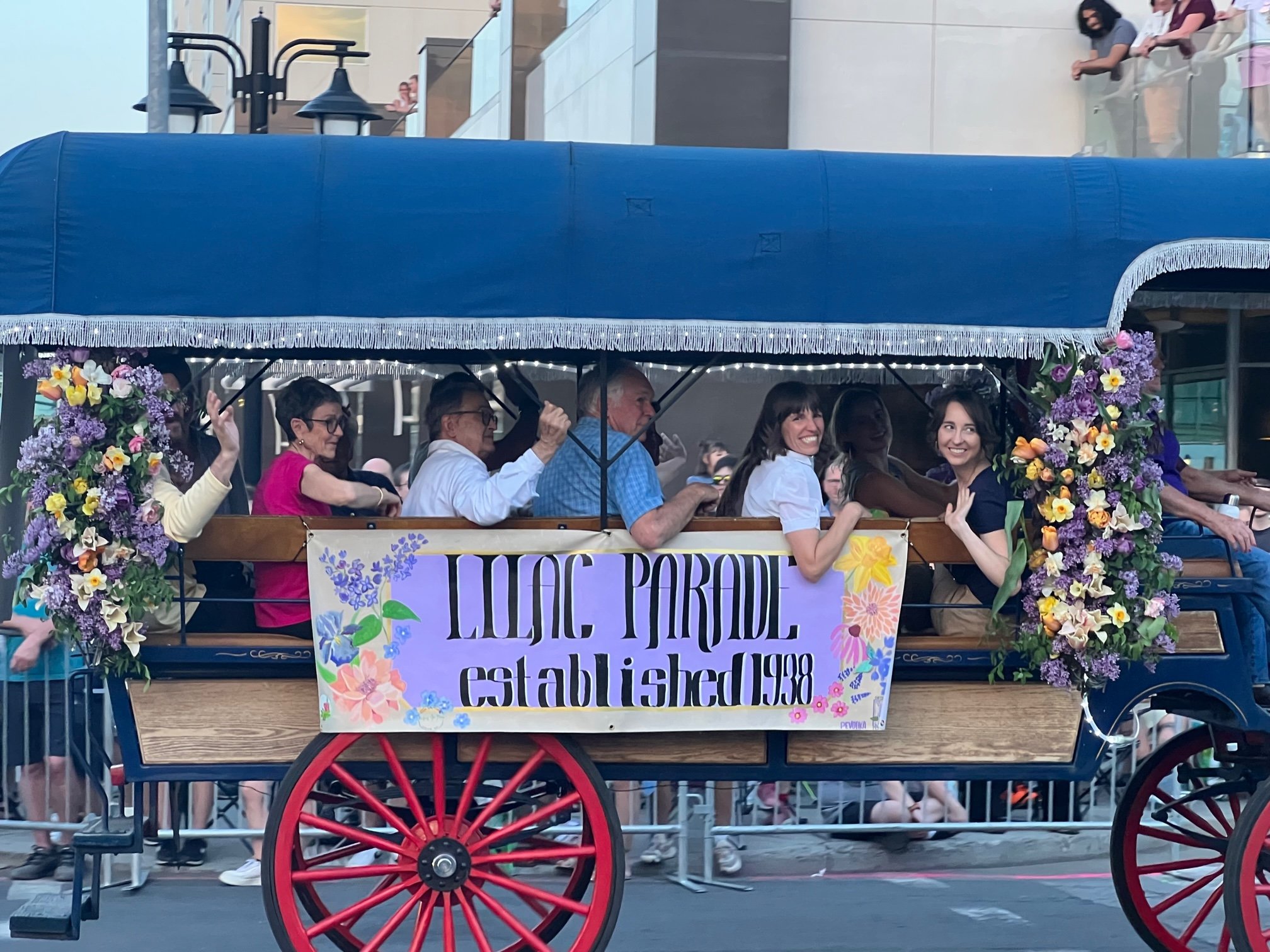 A decorated wagon with a "Lilac Parade Established 1938" sign, carrying several people. The wagon features floral arrangements and is part of a parade, with onlookers in the background.