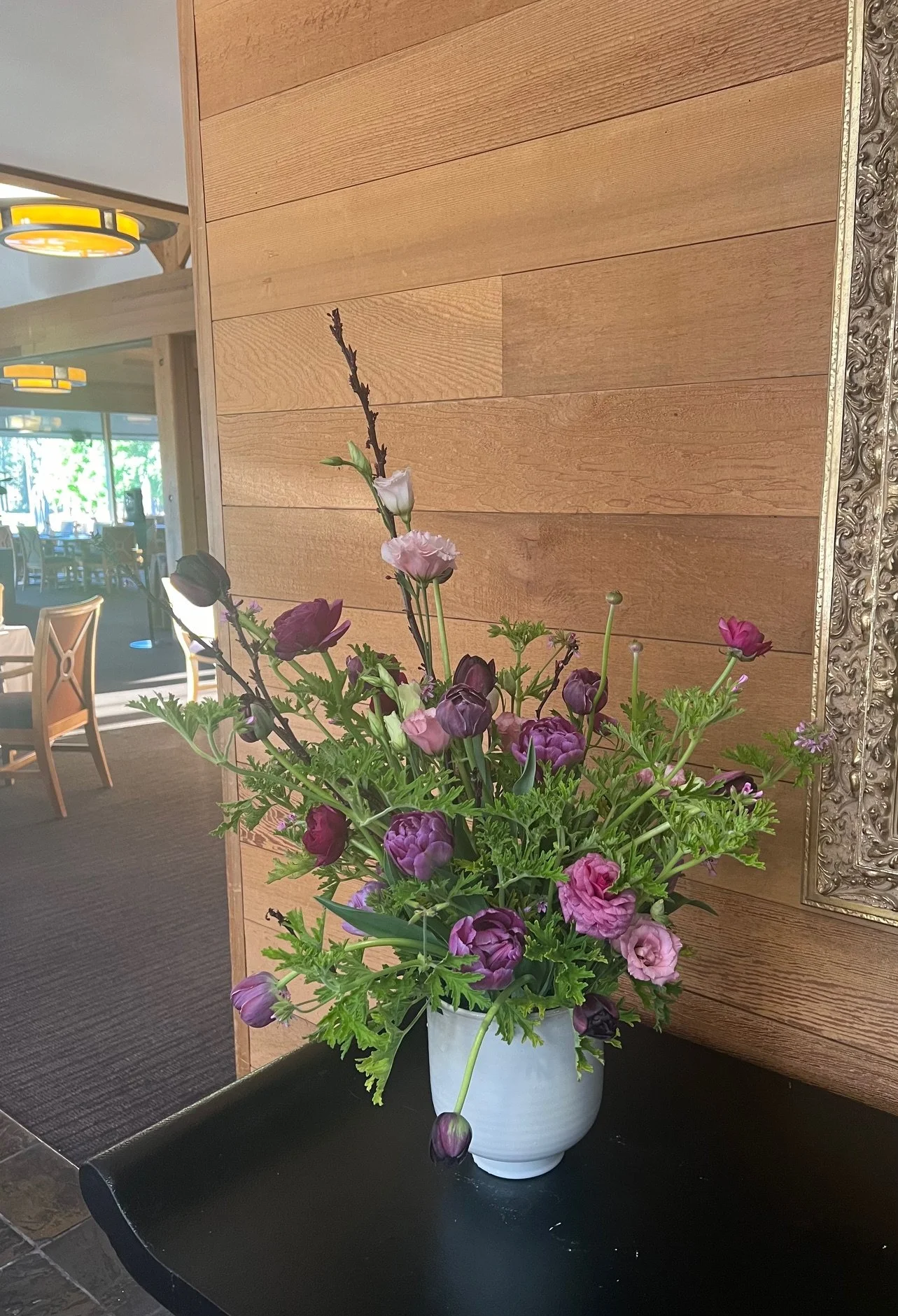 A bouquet of purple and pink flowers with green leaves in a white vase on a dark table against a wooden wall backdrop.