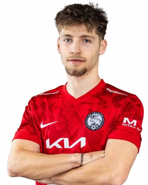 A young man with light brown hair and a beard wearing a red sports jersey with a logo on the chest, crossing his arms.