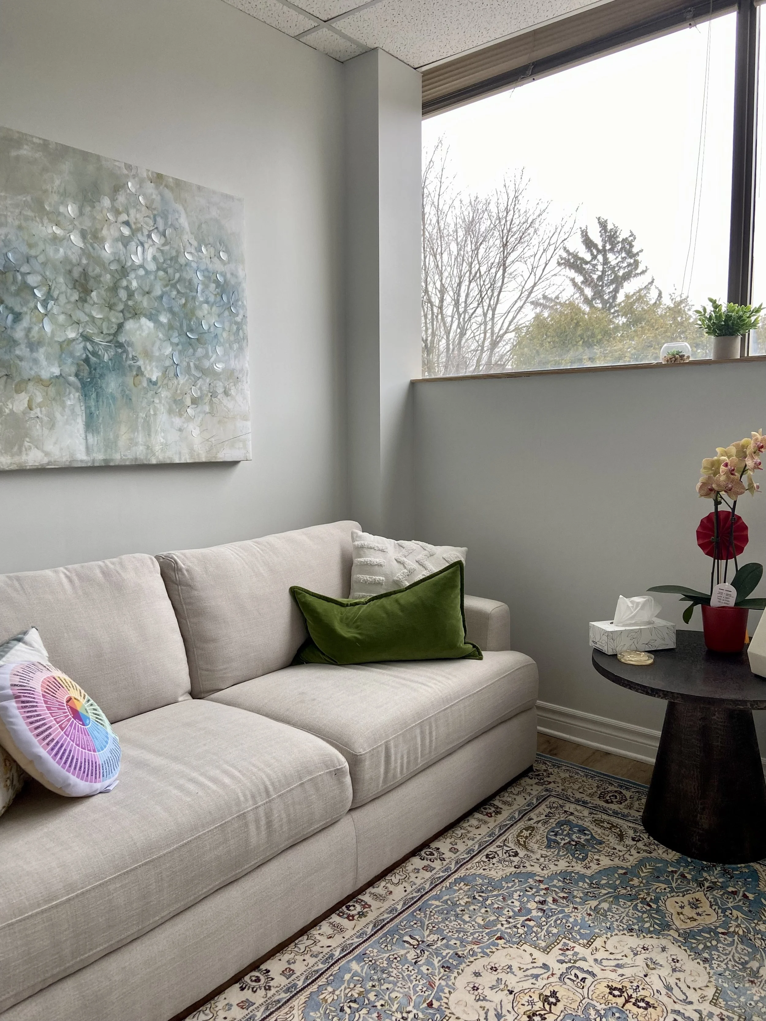 Living room with a beige sofa adorned with colorful and green pillows, a dark round side table with a potted orchid, tissue box, and decorative items, a large window with a view of leafless trees, a patterned rug, and a white wall with a floral painting.