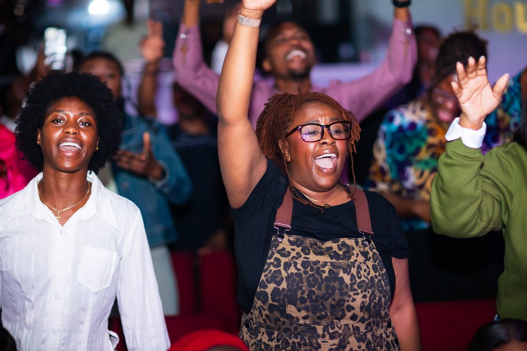 Crowd enjoying a live event, with people raising their hands and singing along enthusiastically. Two women are prominently visible, wearing casual clothes, one in a white shirt and the other in a leopard print outfit.