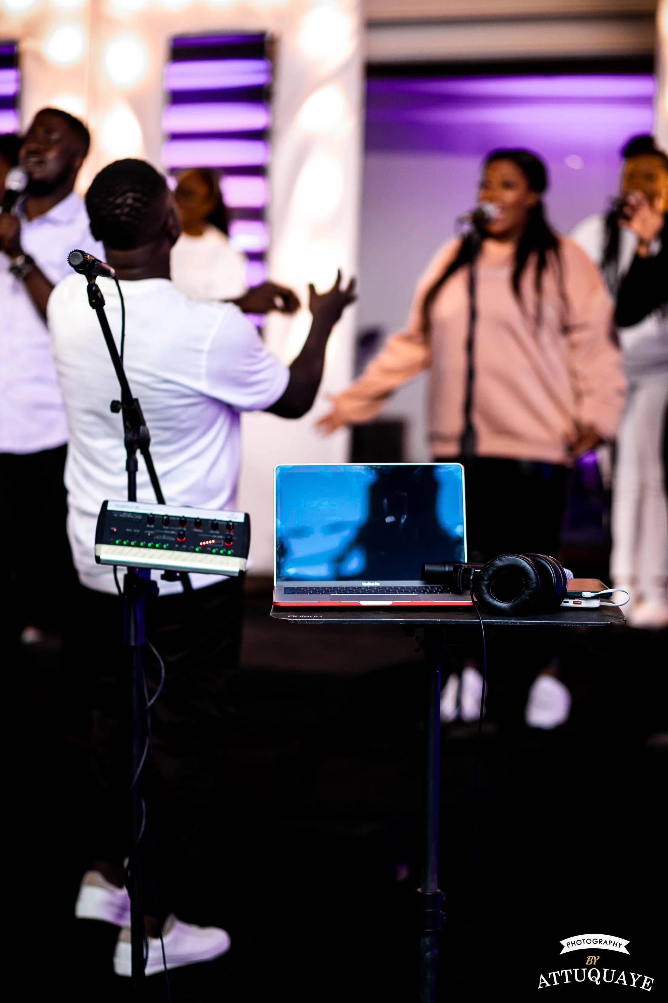 A group of singers performing on stage, with a sound engineer in the foreground. A laptop and headphones are placed on a stand, with a small audio mixer visible next to it. The background is lit with purple and white lights.