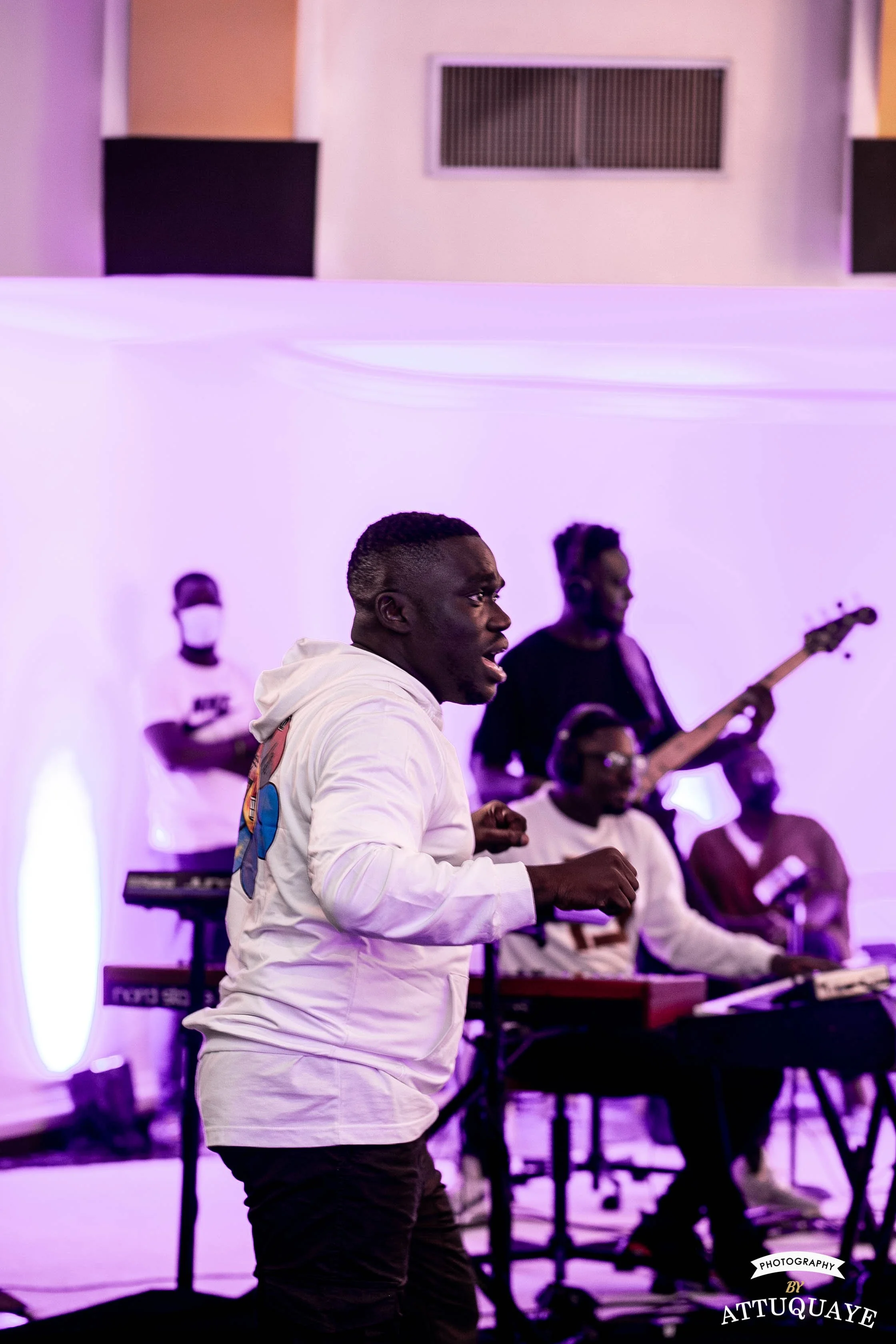 Male musician performing with a band in a purple-lit room, wearing a white hoodie. A bassist and keyboardist are visible in the background.