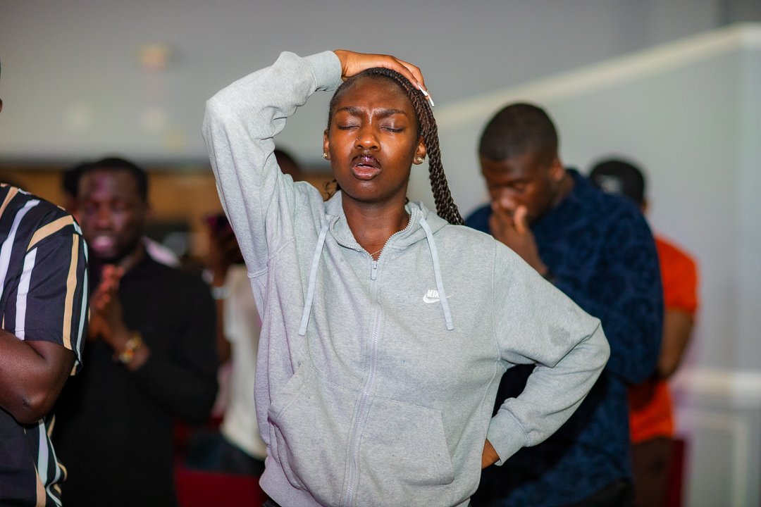 A person wearing a gray hoodie with their eyes closed and hand on their head in a crowded indoor setting.