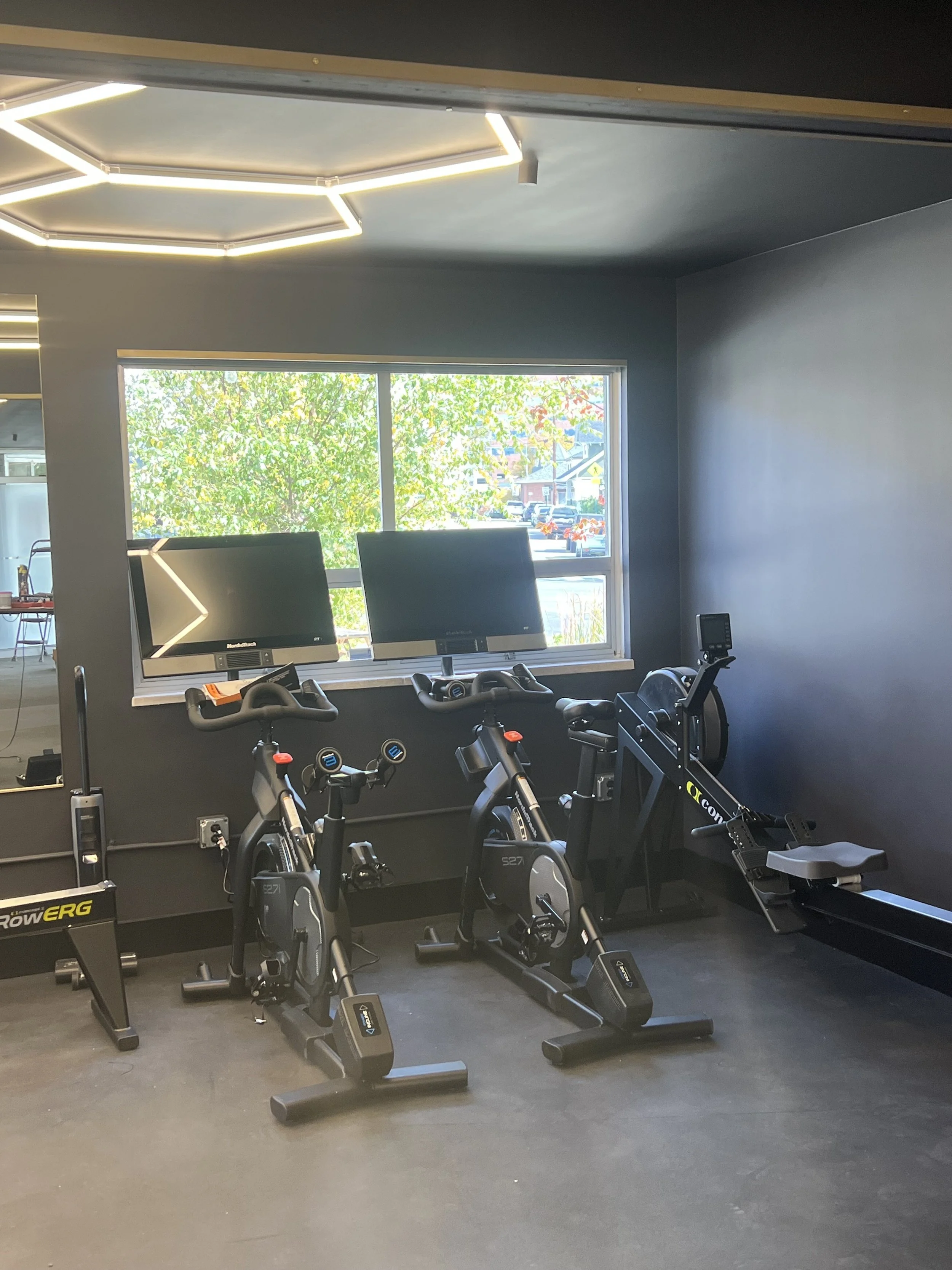 Indoor gym with two stationary bikes and a rowing machine by a window, illuminated by geometric ceiling lights.