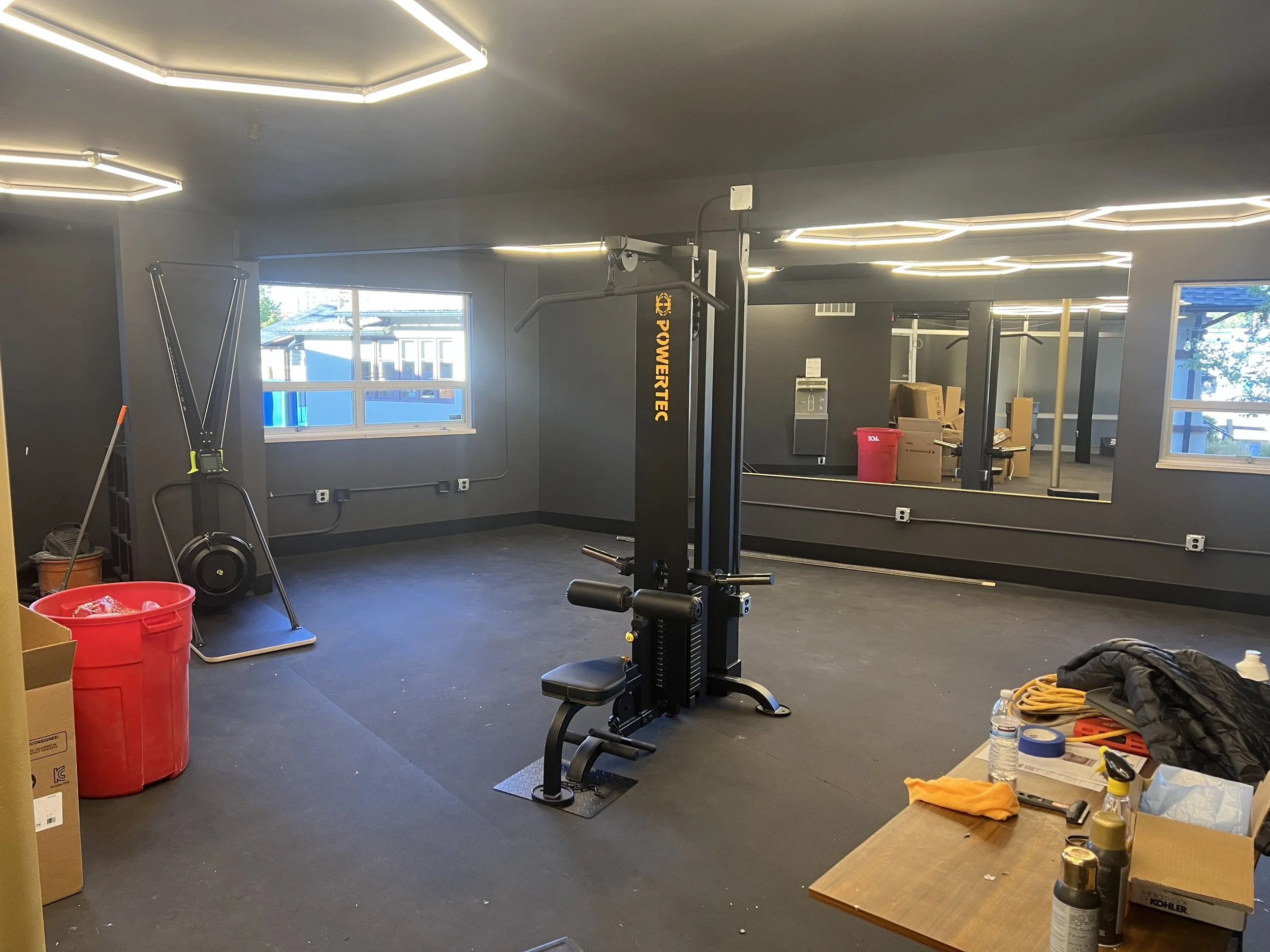 Empty gym with workout equipment including a power tower, weights, and miscellaneous items on a table, with windows letting in natural light.