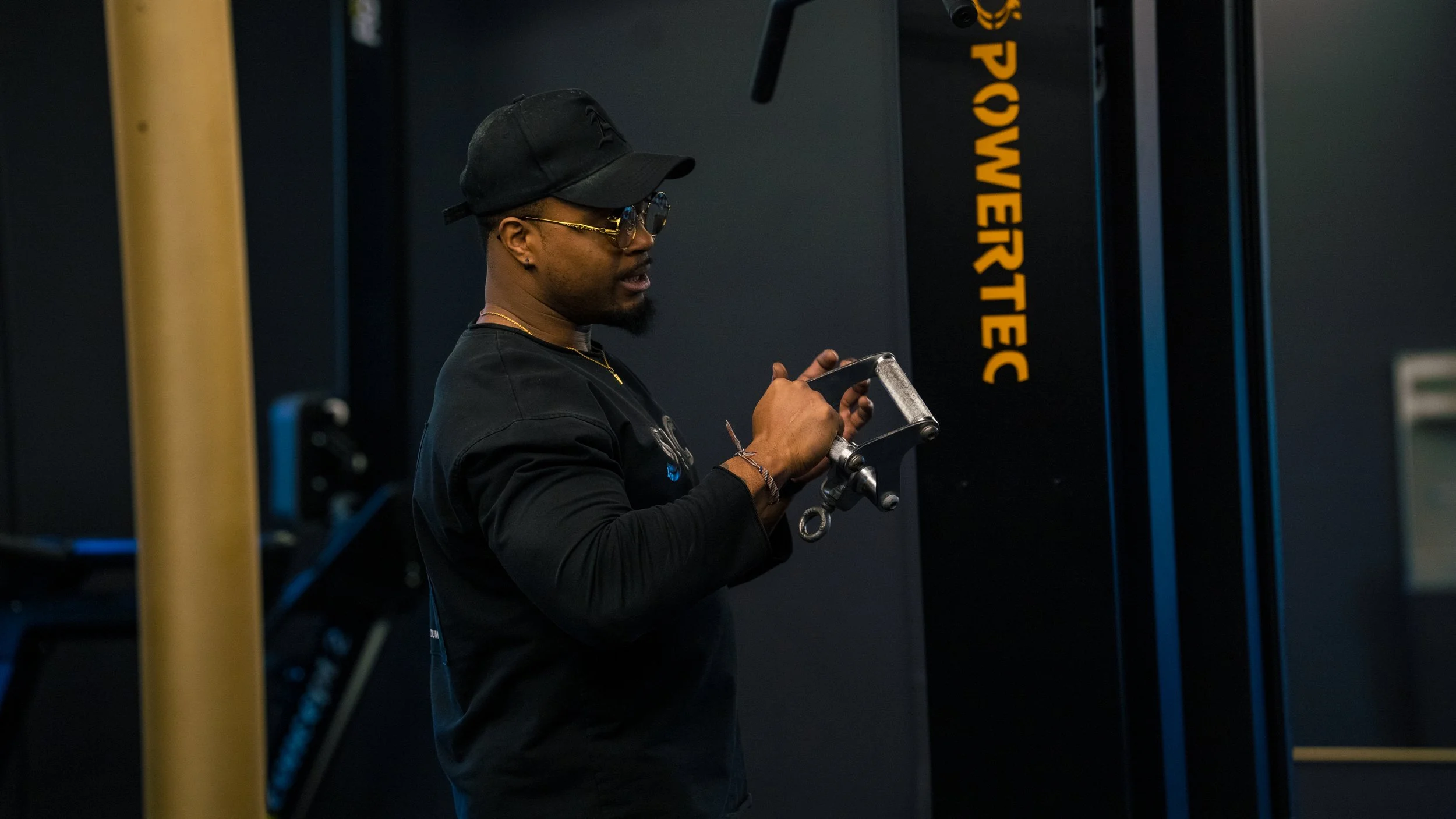 A man wearing a black cap, glasses, and a black long-sleeve shirt is standing in a gym, using his phone with his right hand.