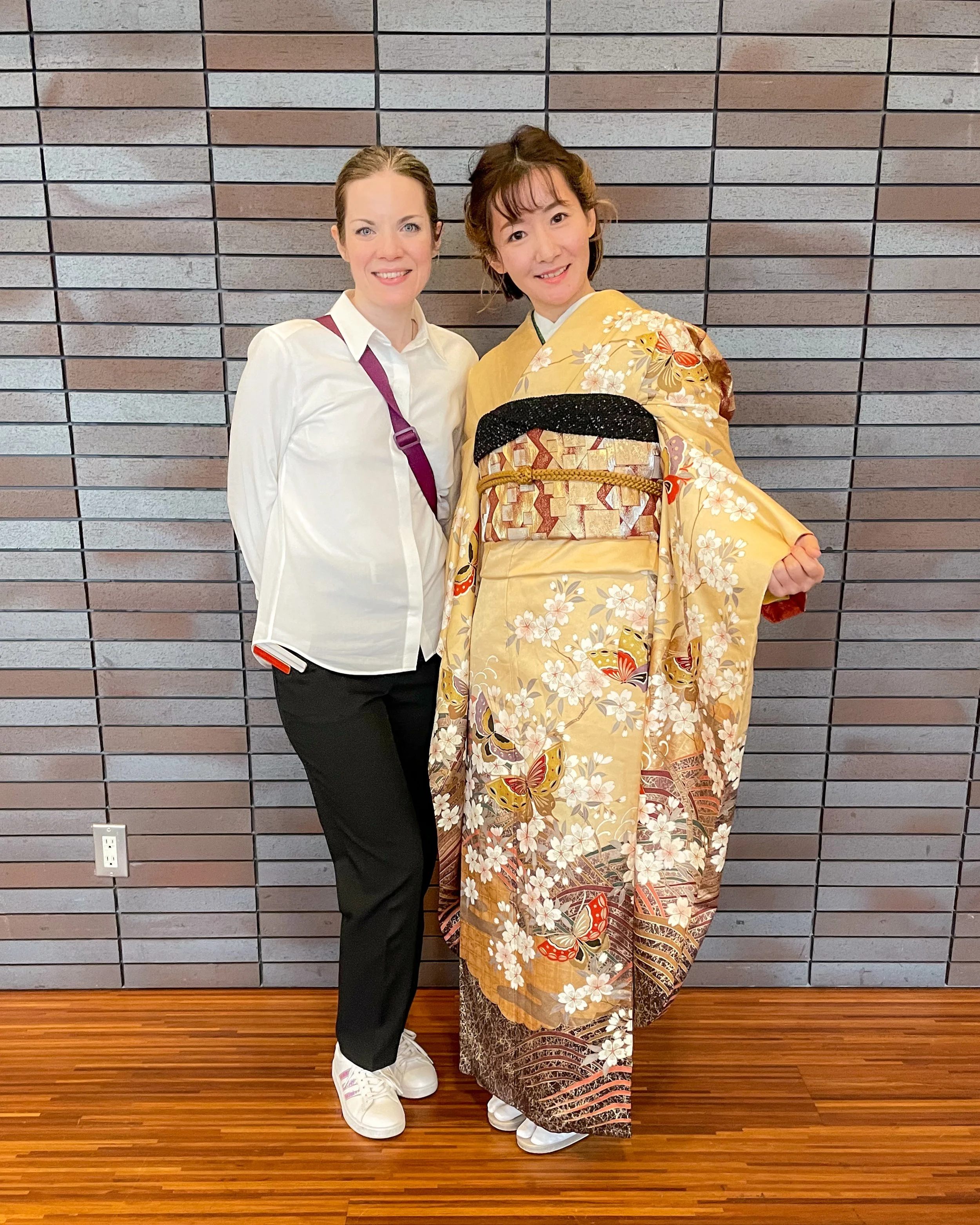Two women standing side by side against a brick wall. One is dressed in a white shirt, black pants, and sneakers with a purple strap. The other is dressed in a traditional yellow kimono with floral and butterfly patterns, a black and gold obi belt, and white footwear.