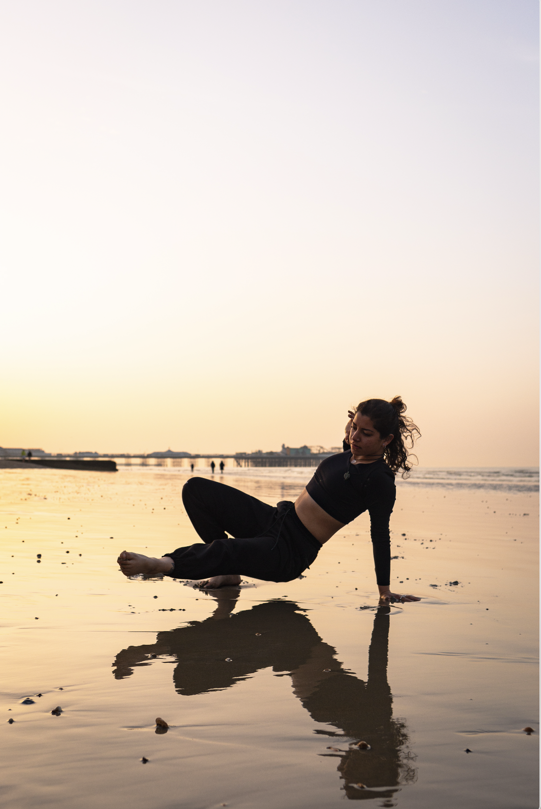 A woman in black athletic clothing is sitting on a wet beach at sunset, with her legs stretched out and leaning slightly back on her arm, near the shoreline, with her reflection visible on the wet sand.