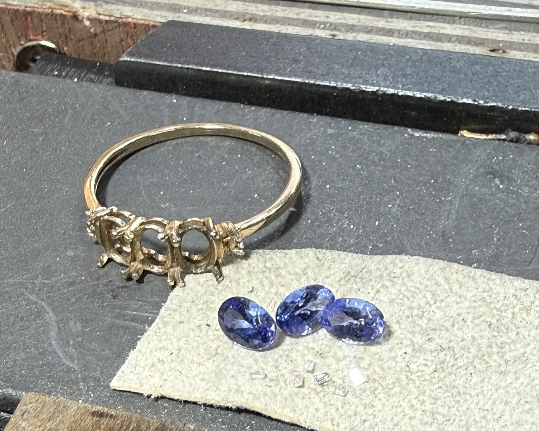 Gold-colored ring with multiple interconnected loops, resting on a textured surface. Below the ring, there are three large, faceted, deep blue gemstones placed on a gray section of the surface. The background features black and gray textured surfaces, with some wooden elements visible at the top of the image.