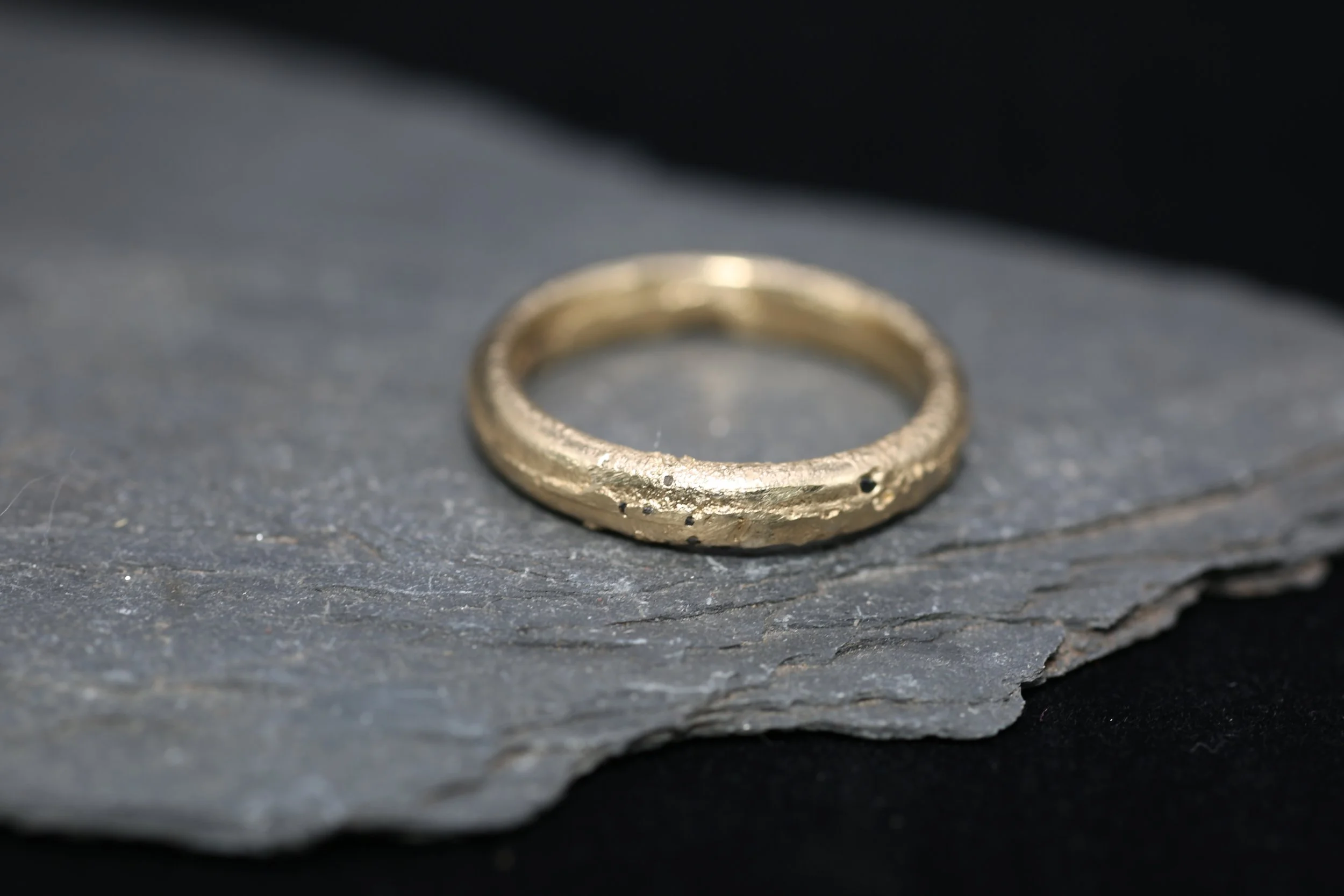 Close-up of a gold ring with a textured surface, resting on dark, textured slate or stone surface.
