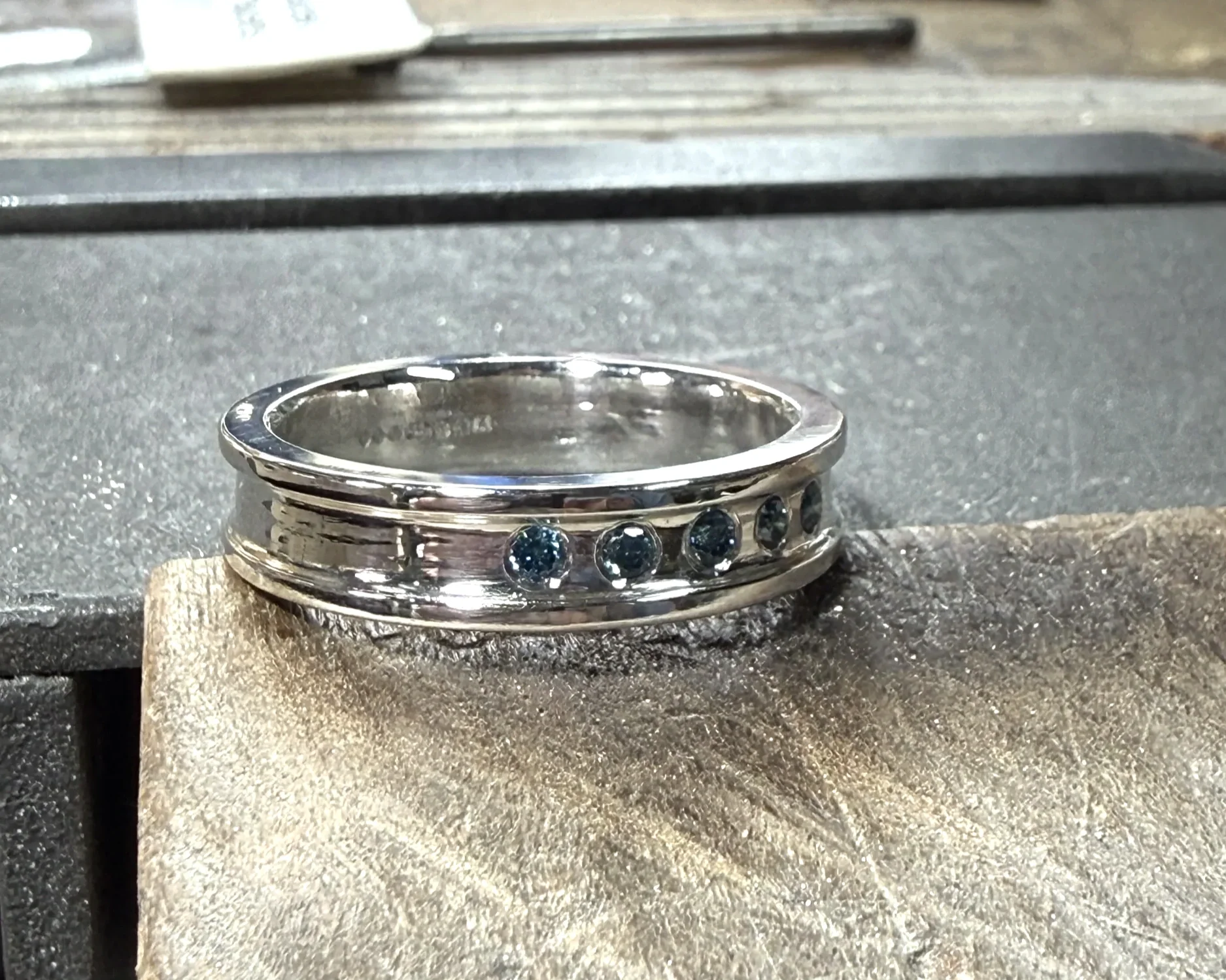 Close-up of a metal ring with blue gemstones set around its circumference, placed on a workbench in a jewelry workshop.