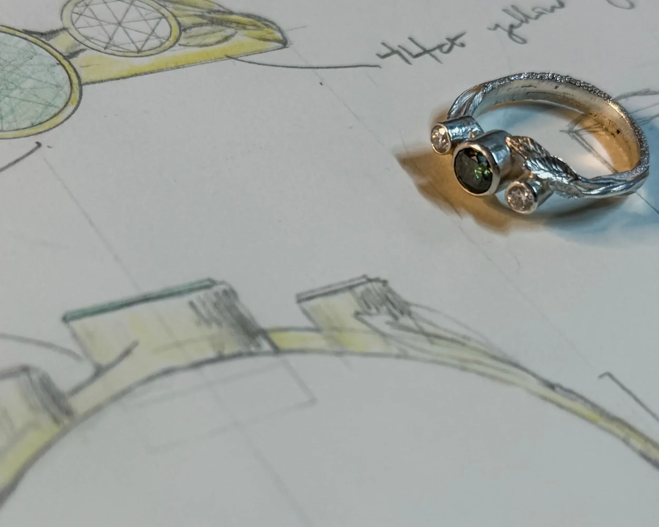 Close-up photo of a shiny silver ring with diamonds resting on a piece of paper with hand-drawn sketches of glasses in yellow and black ink.