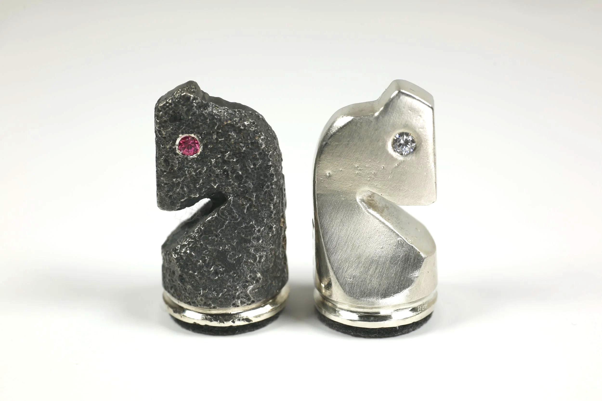 Two metal objects shaped like the letter 'K', one black with a textured surface and a pink gemstone eye, the other silver with a smooth surface and a clear gemstone eye.