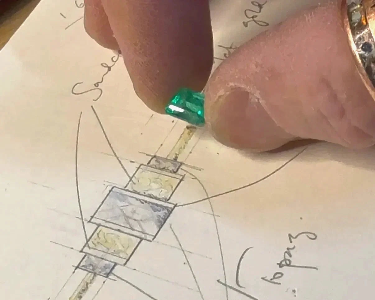 Iain Sainsbury measuring an emerald on a hand-drawn jewellery design sketch with labeled notes and measurements.